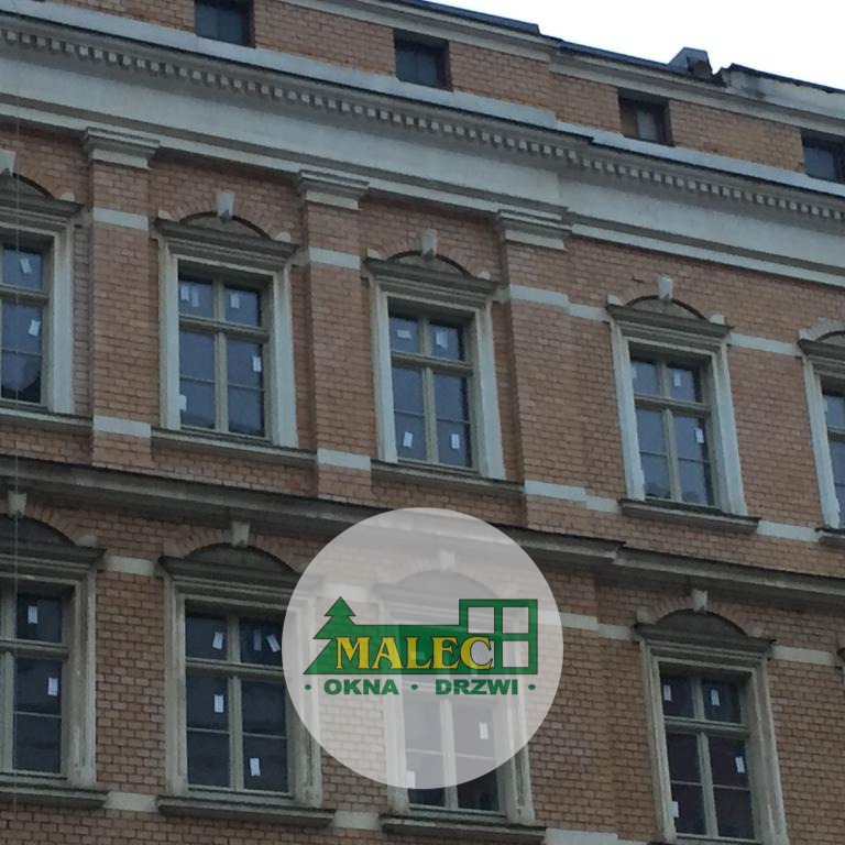 Fasada kamienicy z oknami drewnianymi, częściowo zasłonięta logo firmy Malec Okna Drzwi w okrągłej ramce.