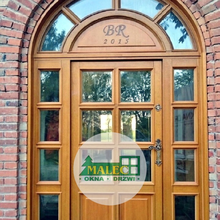 Drewniane drzwi wejściowe z łukowym nadświetlem i bocznymi przeszkleniami, umieszczone w murze z czerwonej cegły. Nadświetle zdobione napisem 'BR 2015'. Na drzwiach widoczne logo firmy.