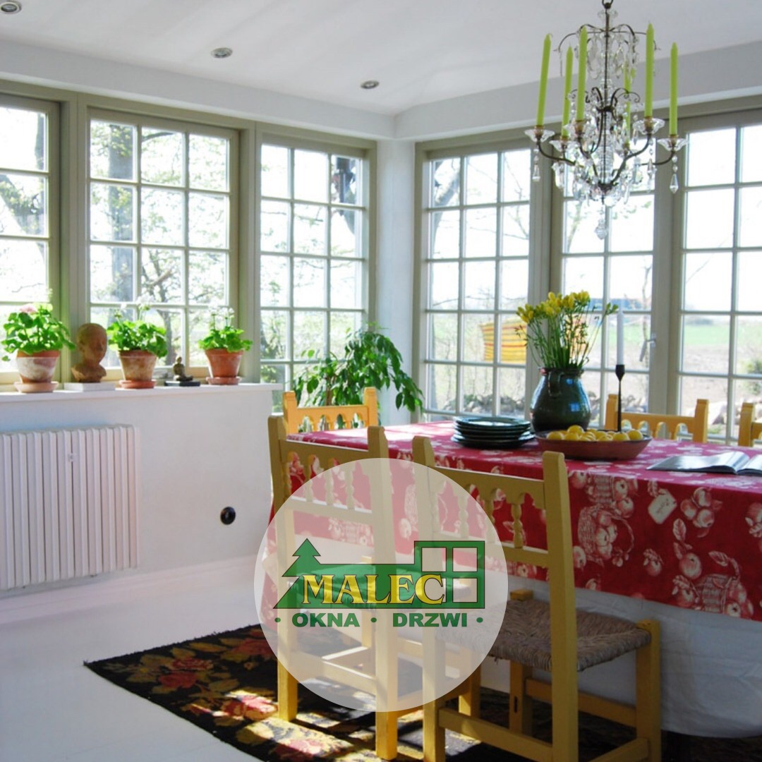 Jasna jadalnia z dużymi, wielokwaterowymi oknami, roślinami na parapecie i ozdobnym żyrandolem, logo firmy na pierwszym planie.
