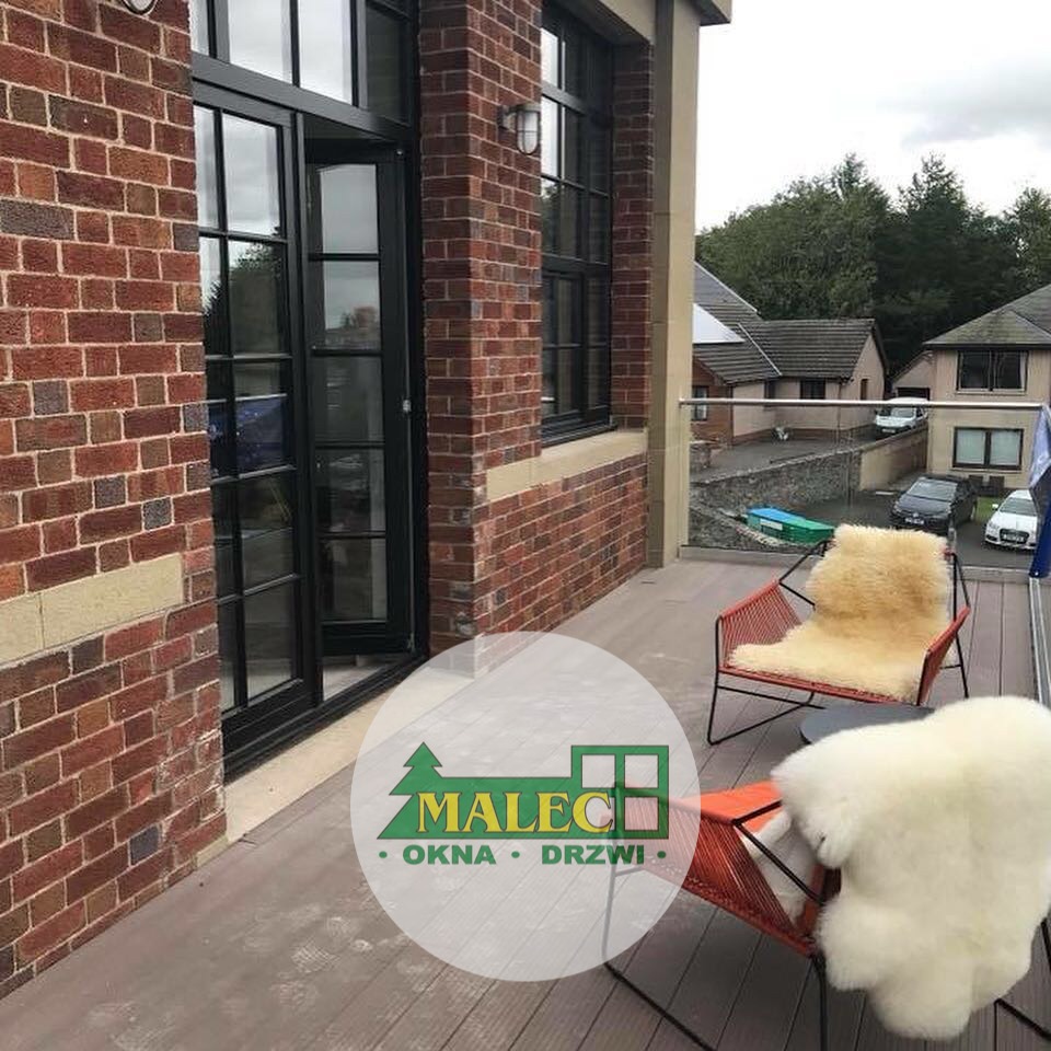 Czarne, wielokwaterowe drzwi balkonowe otwarte na taras z ceglaną ścianą i krzesłami z futrzanymi narzutami, widok na domy w tle.