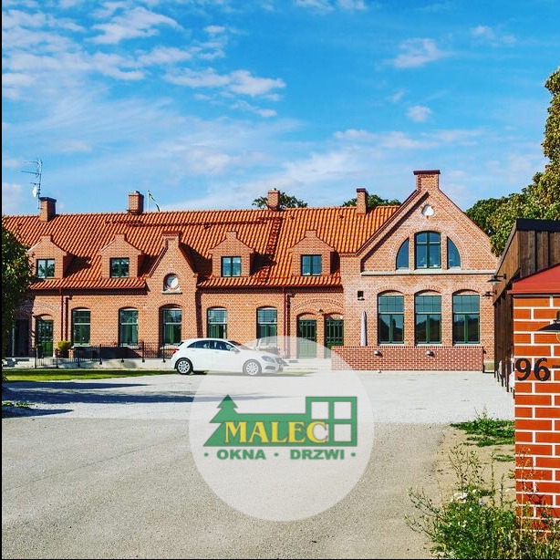 Ceglany budynek z czerwonym dachem i ciemnozielonymi ramami okiennymi, biały samochód zaparkowany przed budynkiem, logo firmy na pierwszym planie.