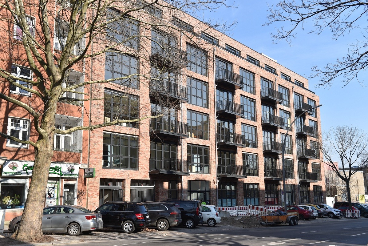 Nowoczesny budynek z cegły z dużymi oknami i balkonami, widok z ulicy z zaparkowanymi samochodami i drzewem na pierwszym planie.