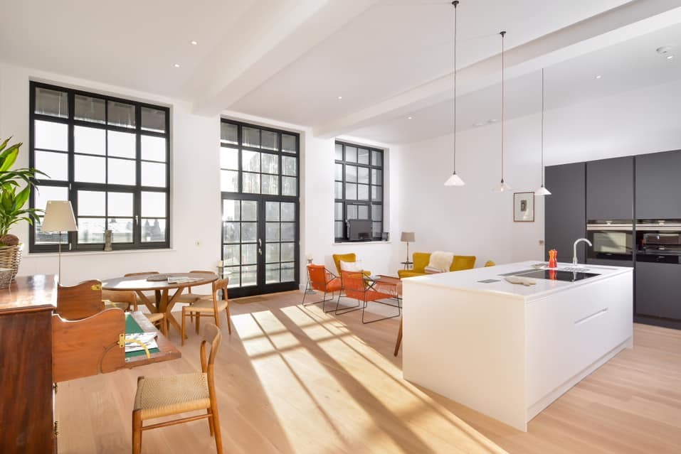 Jasne wnętrze z dużymi, czarnymi oknami i drzwiami balkonowymi. Widoczne meble w stylu retro i nowoczesna wyspa kuchenna. Promienie słoneczne wpadają do środka.