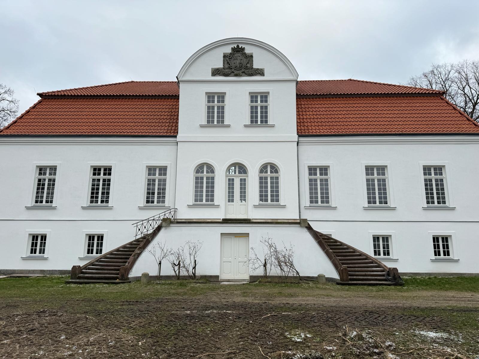 Biały, dwukondygnacyjny budynek z czerwonym dachem, symetrycznymi schodami i oknami. Widoczne detale architektoniczne i elementy dekoracyjne na fasadzie.