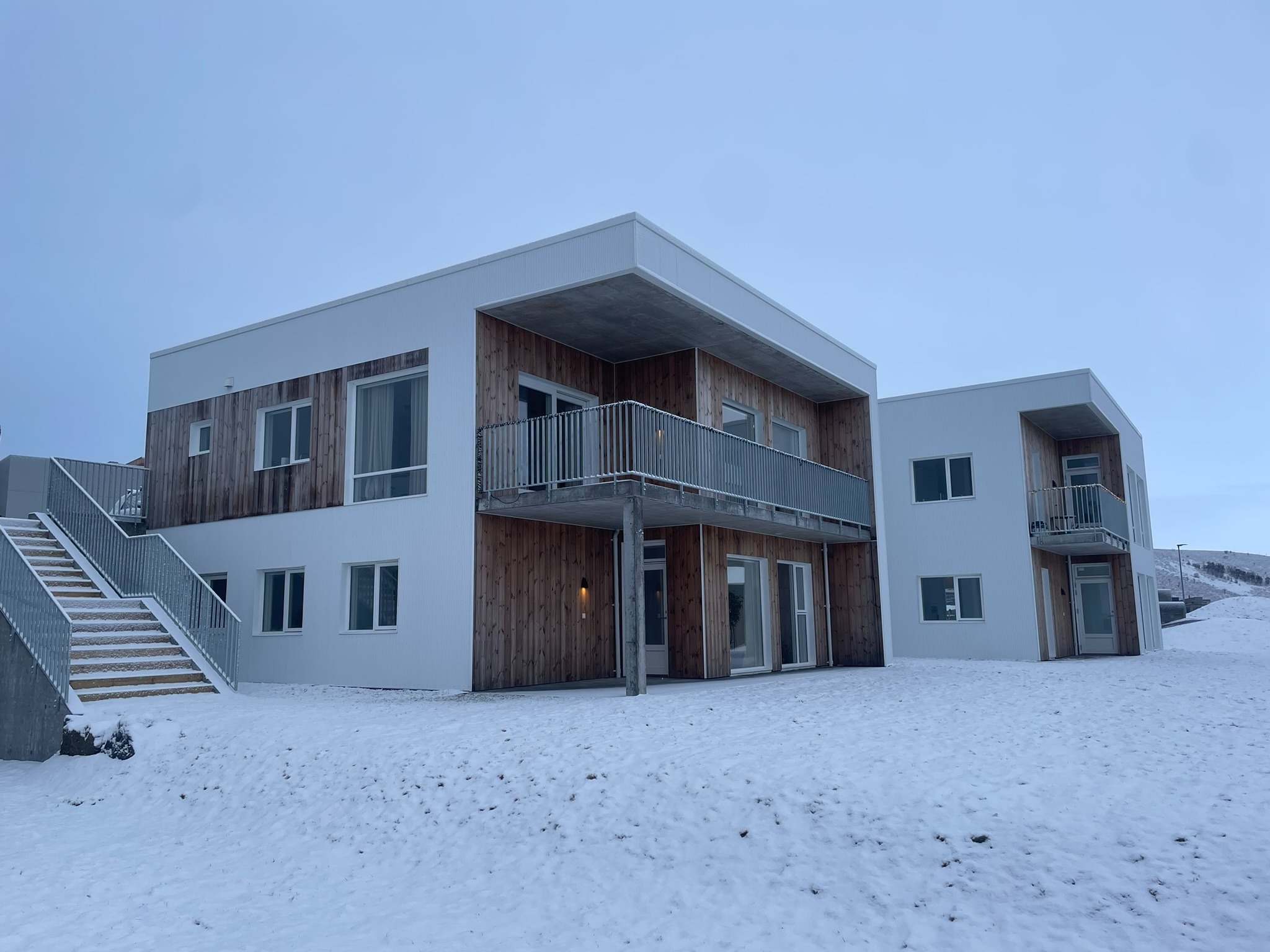 Nowoczesny budynek z drewnianą elewacją i balkonami, zimowy krajobraz. Widoczne schody zewnętrzne i okna. Minimalistyczny design w skandynawskim stylu.