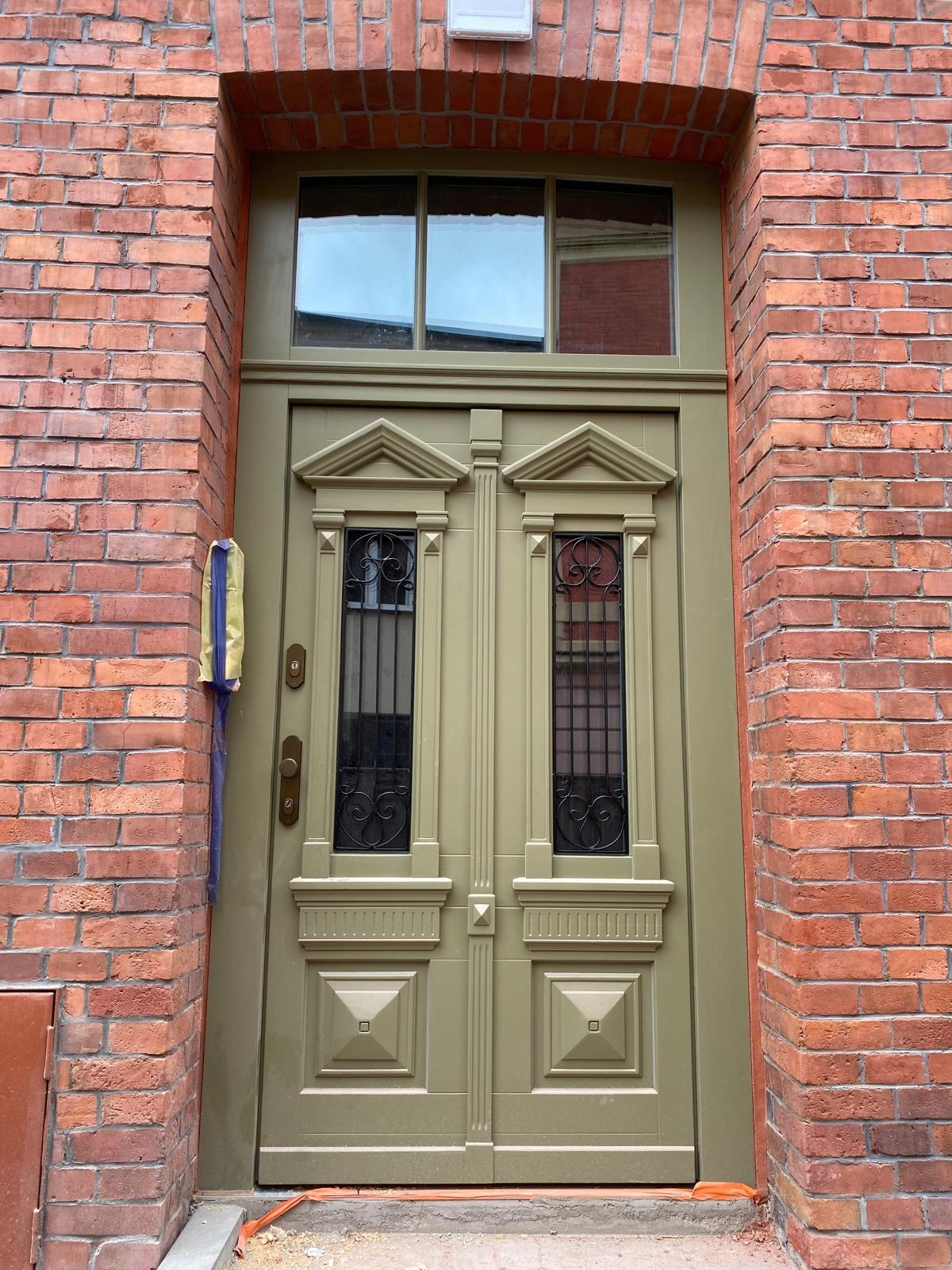 Eleganckie, dwuskrzydłowe drzwi wejściowe w odcieniach oliwkowej zieleni, z ozdobnymi kratami i przeszkleniem nad drzwiami, wkomponowane w czerwoną ceglaną fasadę budynku.