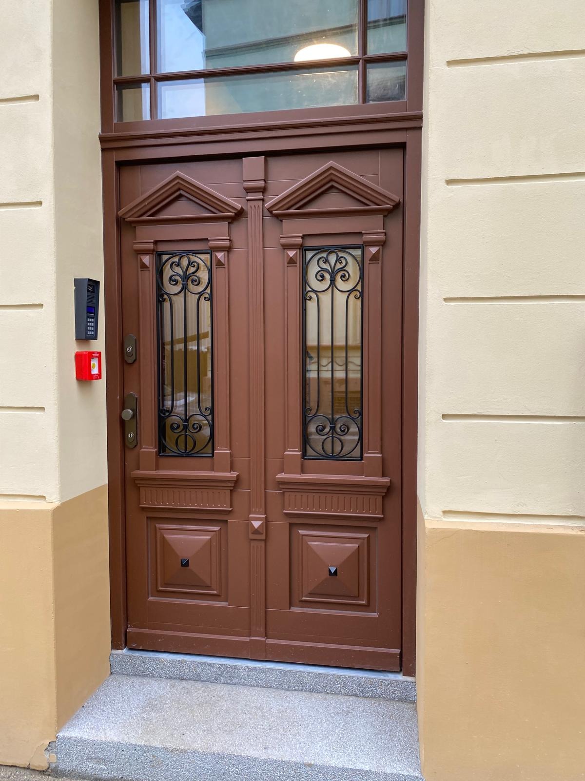 Dwuskrzydłowe, brązowe drzwi wejściowe z ozdobnymi, metalowymi kratami w szybach, zamontowane w budynku z beżową elewacją i domofonem po lewej stronie.