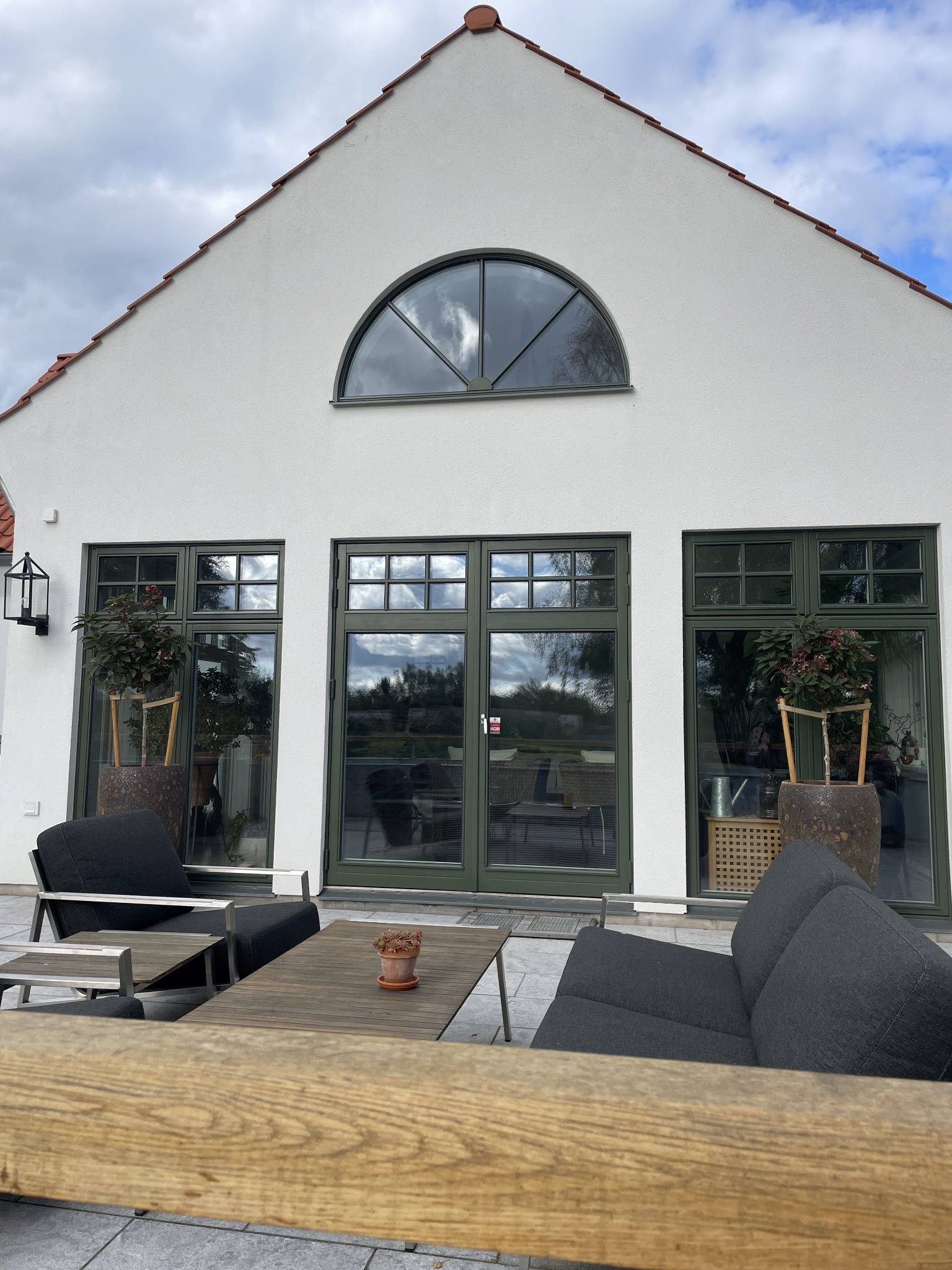 Biały budynek z zielonymi oknami i drzwiami tarasowymi. Półokrągłe okno na szczycie. Meble ogrodowe na tarasie. Widok na zewnątrz przez szyby.