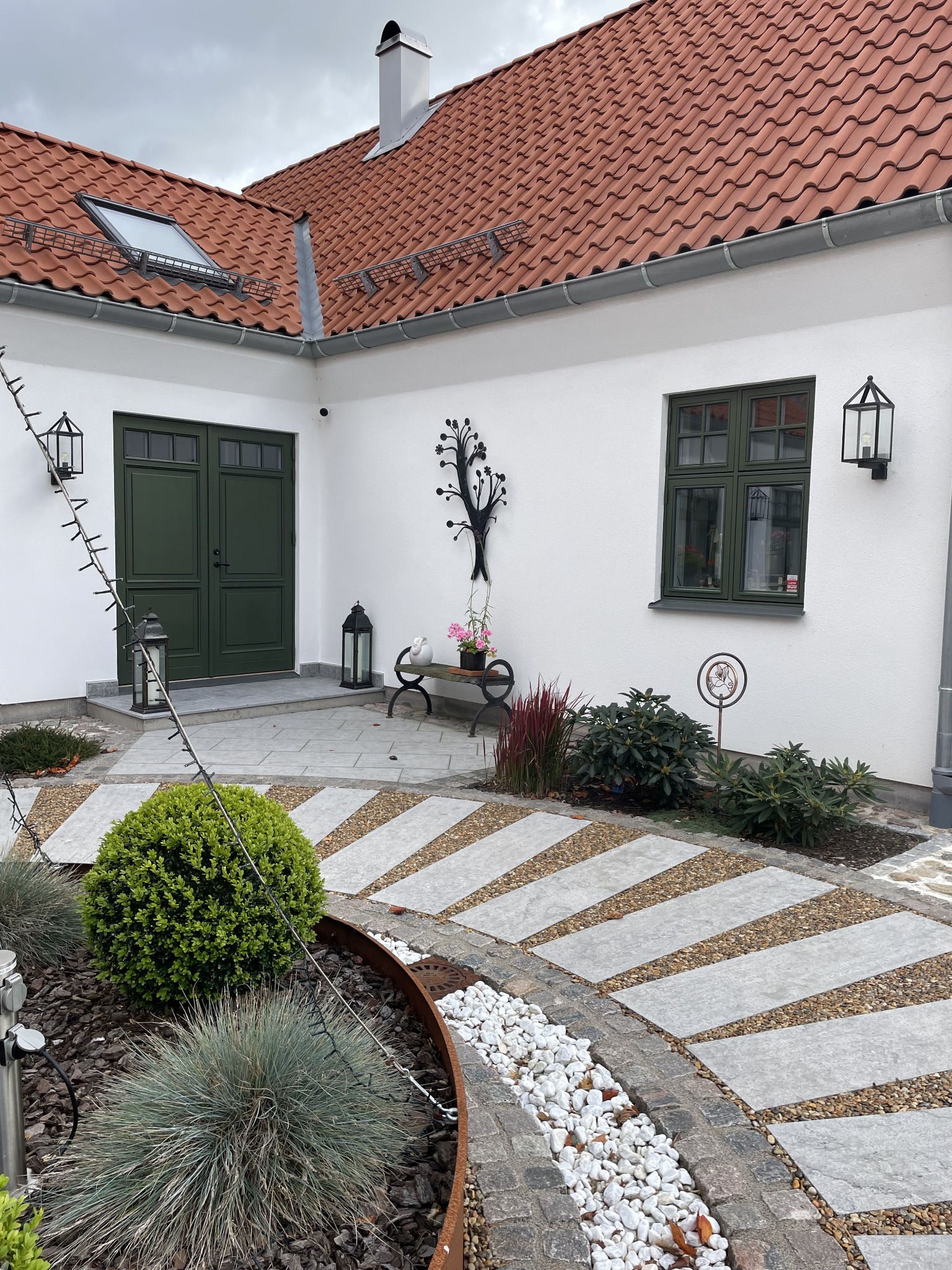 Eleganckie, zielone drzwi wejściowe i okno w białym domu z czerwoną dachówką. Uwagę przykuwa starannie zaprojektowany ogród z kamiennymi ścieżkami i dekoracjami.