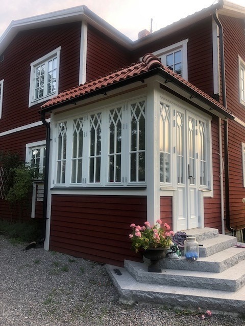 Zadaszone wejście do domu z białymi oknami w stylu gotyckim i dwuskrzydłowymi drzwiami. Czerwona elewacja, kamienne schody i kwiaty w donicy.