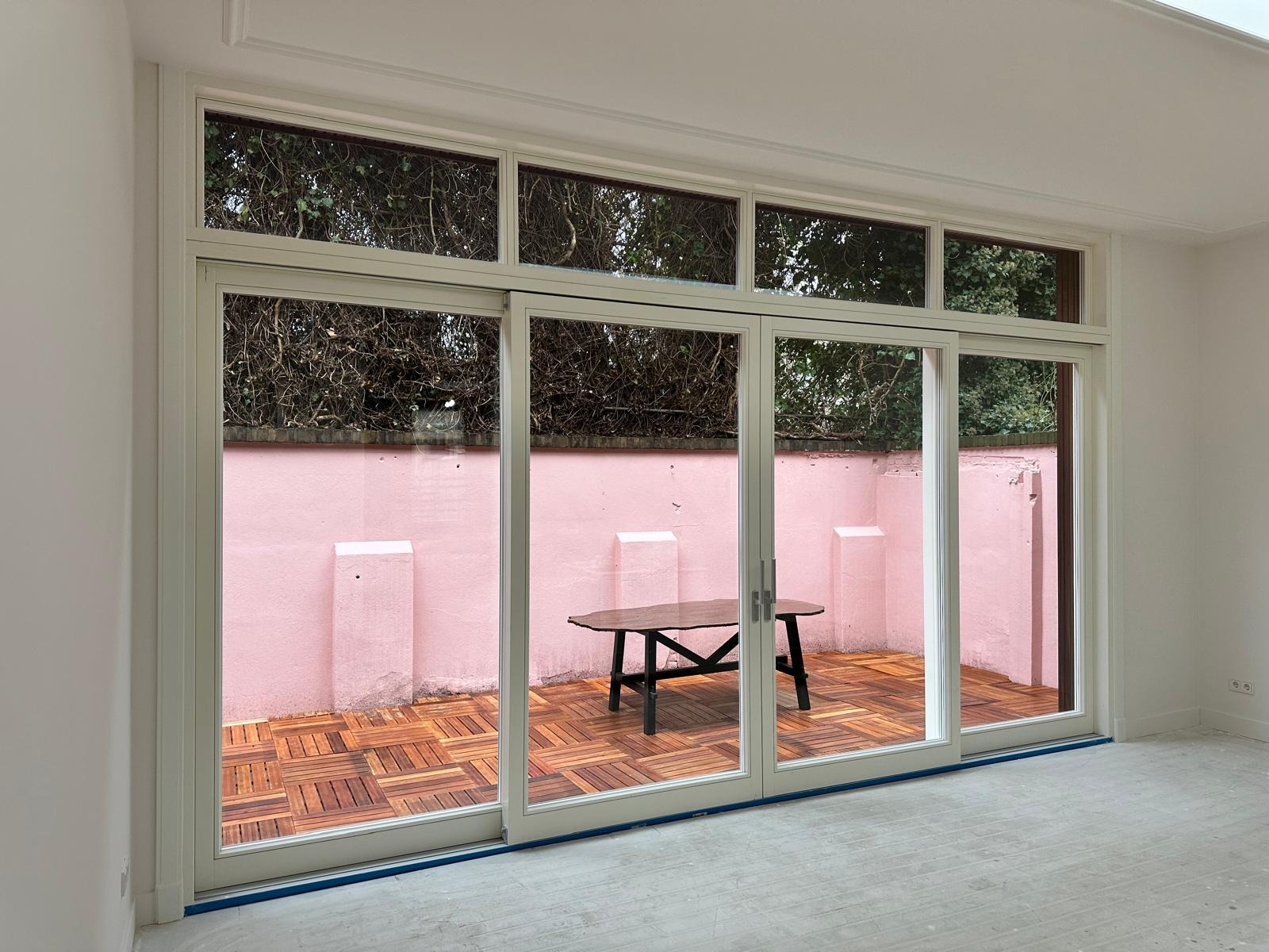 Jasne, trzyskrzydłowe drzwi przesuwne z widokiem na różowy taras z drewnianą podłogą. Minimalistyczny design, naturalne światło, nowoczesne wykończenie wnętrza.