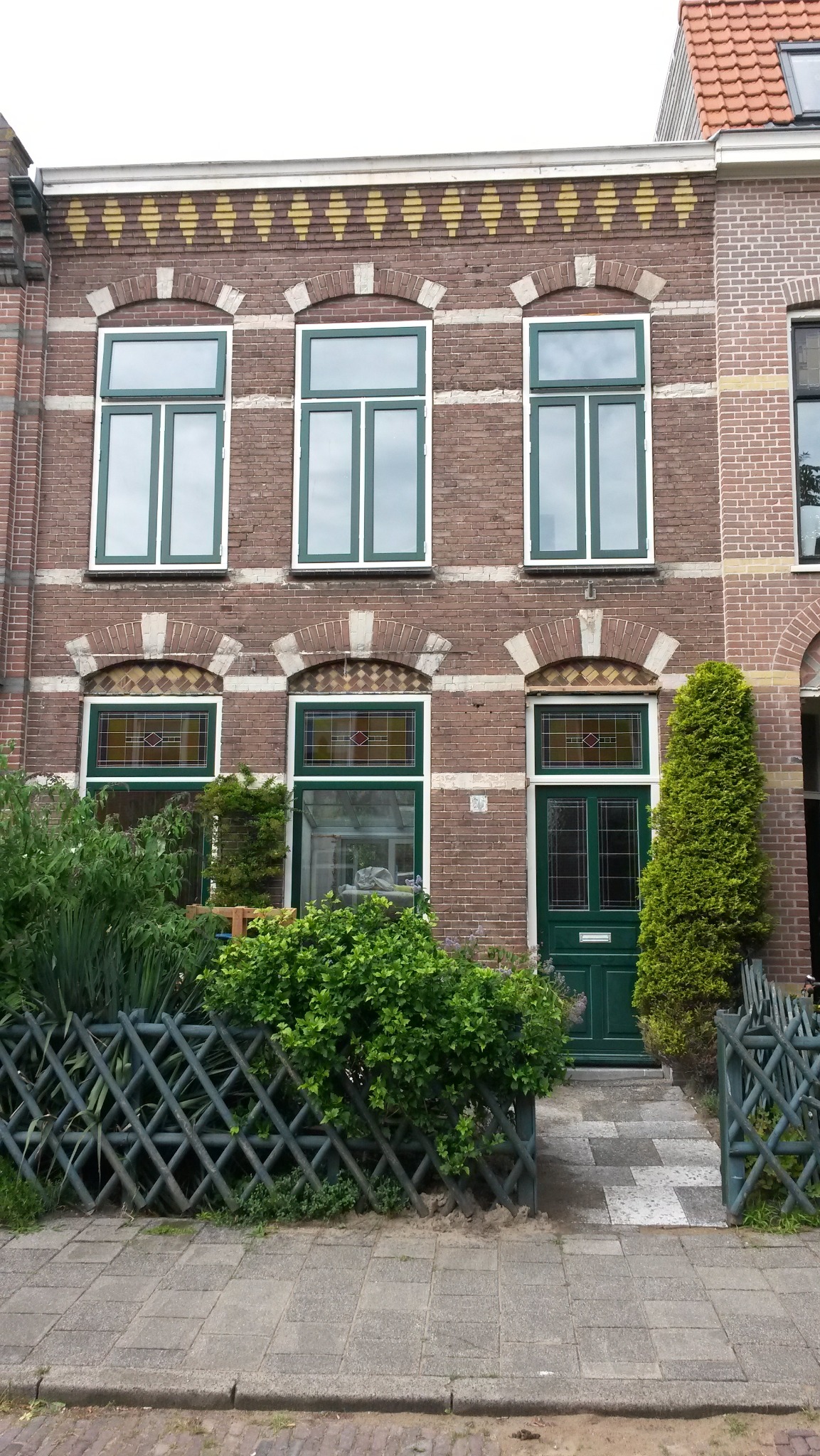 Elewacja budynku z cegły z zielonymi oknami i drzwiami. Nad oknami dekoracyjne łuki z cegły. Przed budynkiem zielony płot i roślinność.