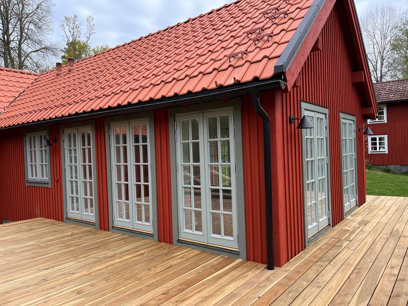 Czerwony dom z drewnianą elewacją i jasnymi, przeszklonymi drzwiami tarasowymi. Widoczny fragment tarasu z desek i dach z czerwonej dachówki.