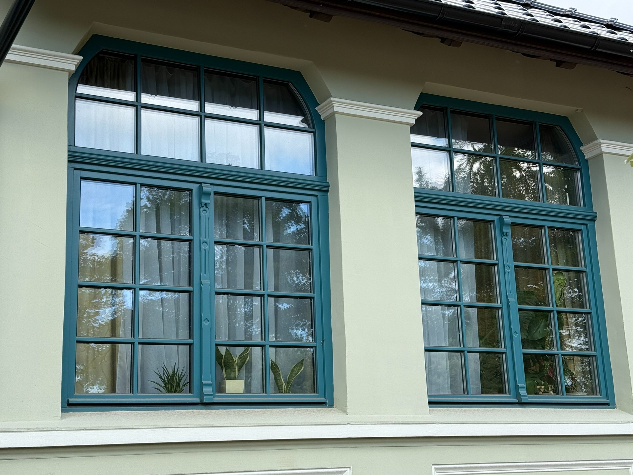 Dwa okna drewniane w kolorze morskim z dekoracyjnymi szprosami i łukowym wykończeniem, widoczne rośliny doniczkowe wewnątrz, jasna elewacja budynku.