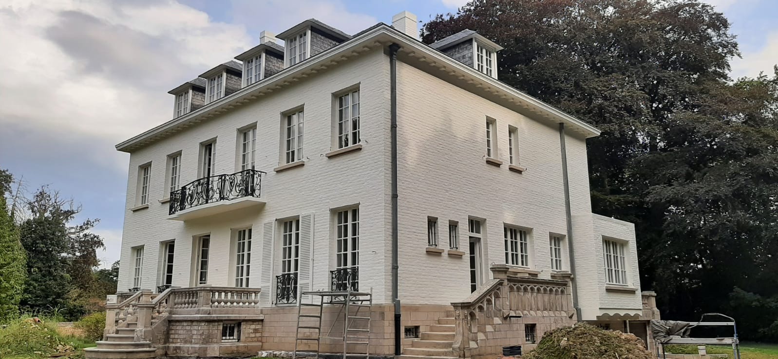 Elewacja dwupiętrowego, białego domu z oknami i balkonem z kutą balustradą. Widoczne rusztowania i prace wykończeniowe. W tle drzewa i pochmurne niebo.