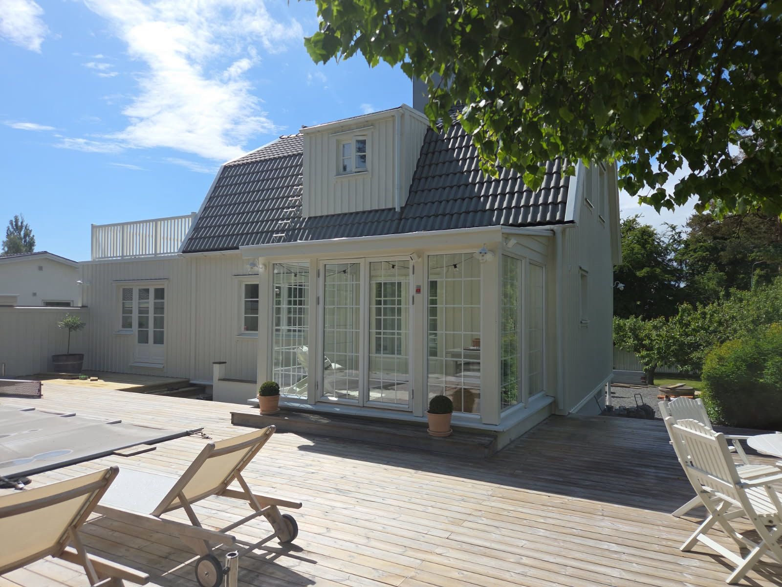 Jasny, skandynawski dom z przeszkloną werandą i tarasem z leżakami. Widok z zewnątrz, słoneczny dzień, błękitne niebo. Charakterystyczna architektura.