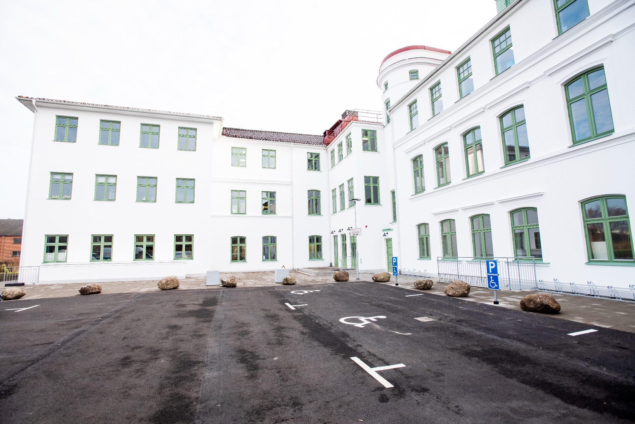 Biały budynek z zielonymi oknami widziany z parkingu dla niepełnosprawnych. Kamienne bariery i świeżo pomalowane pasy na asfalcie.
