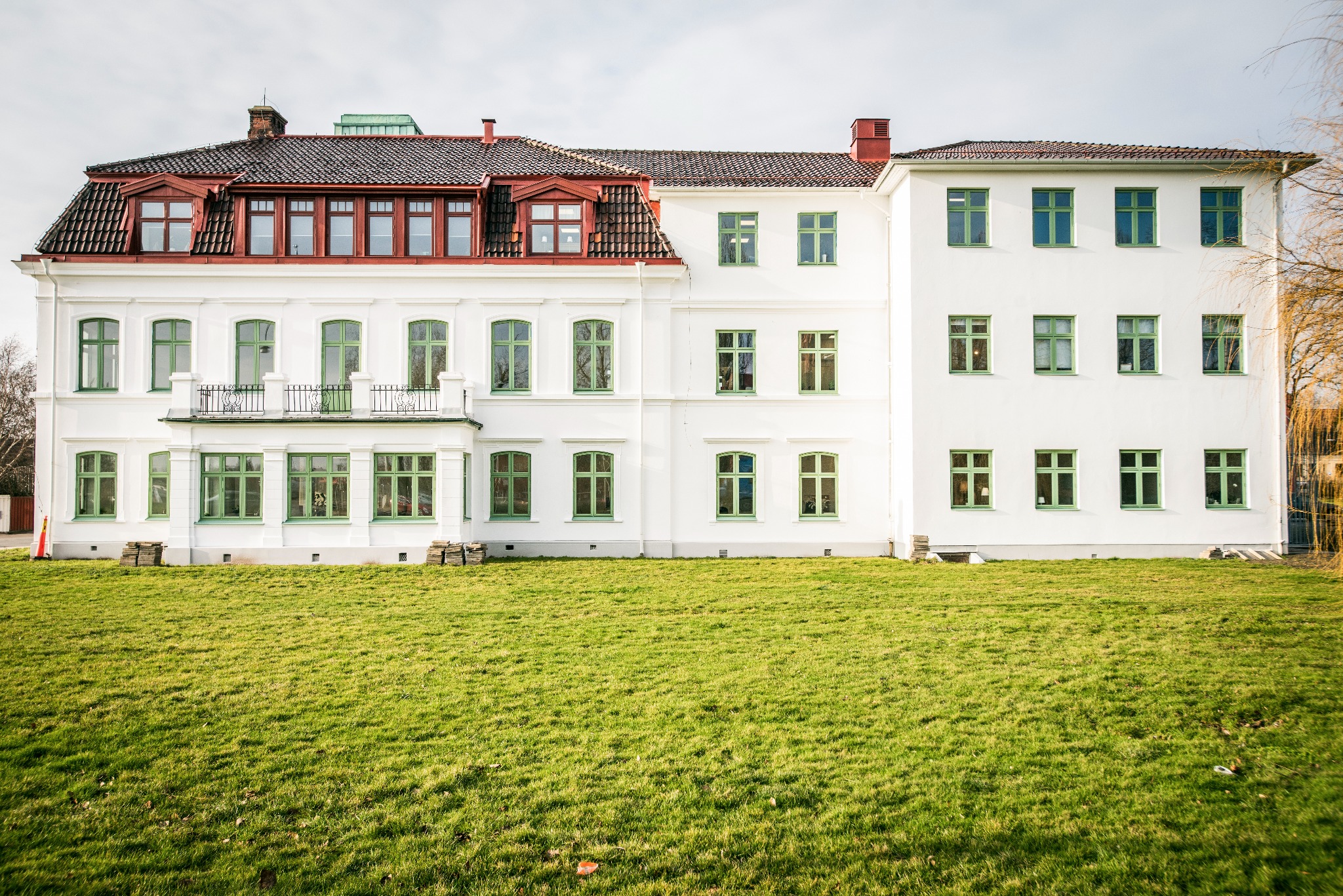 Biały budynek z zielonymi oknami i czerwonym dachem, otoczony trawnikiem. Widoczne detale architektoniczne i naturalne oświetlenie.