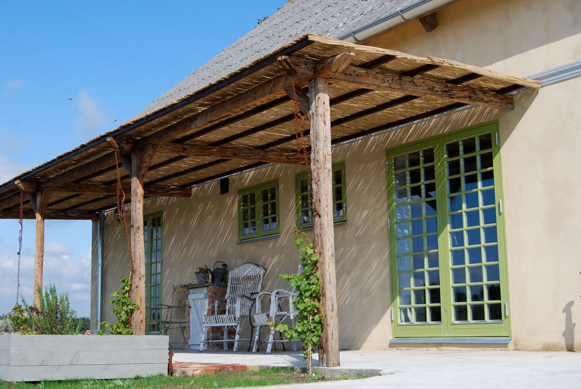 Zadaszony taras z drewnianą konstrukcją i zielonymi oknami oraz dwuskrzydłowymi drzwiami. W tle jasna elewacja budynku i fragment dachu. Uroku dodają rośliny i meble ogrodowe.