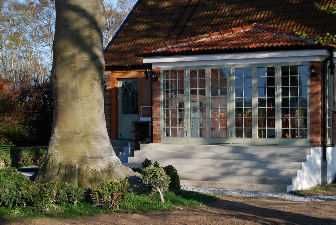 Eleganckie, szklane drzwi harmonijkowe w jasnej, zielonej ramie, prowadzące do domu z cegły. Widoczne schody wejściowe i fragment pnia drzewa.