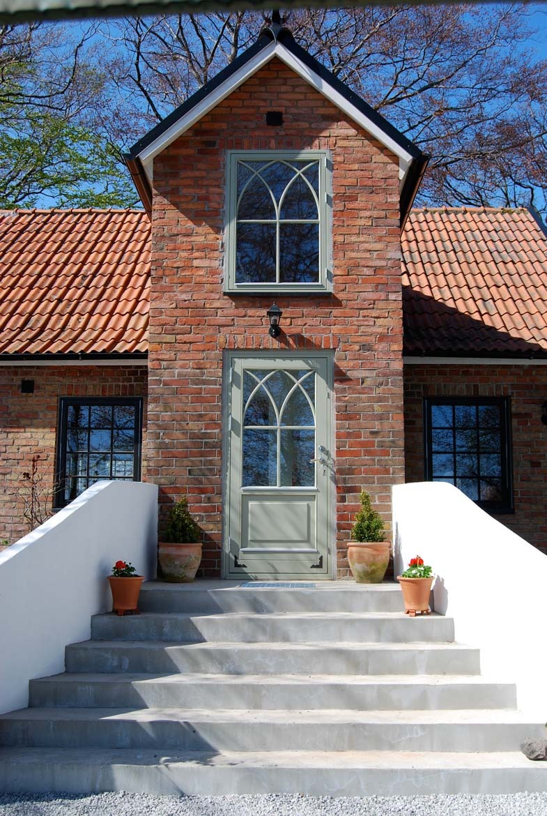 Elewacja domu z cegły z szarozielonymi drzwiami wejściowymi z łukowatym przeszkleniem i oknem na piętrze. Schody prowadzą do wejścia, po bokach donice z roślinami.