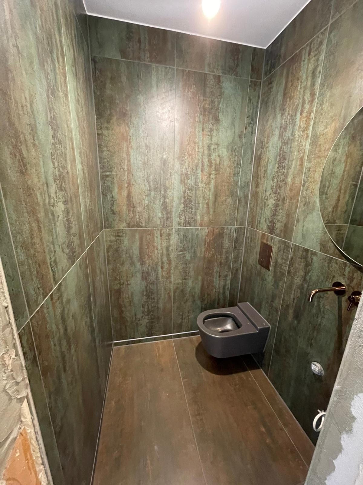 Nowoczesna łazienka z płytkami imitującymi rdzę na ścianach i podłodze, wisząca toaleta w kolorze antracytowym, okrągłe lustro i bateria w odcieniach brązu.