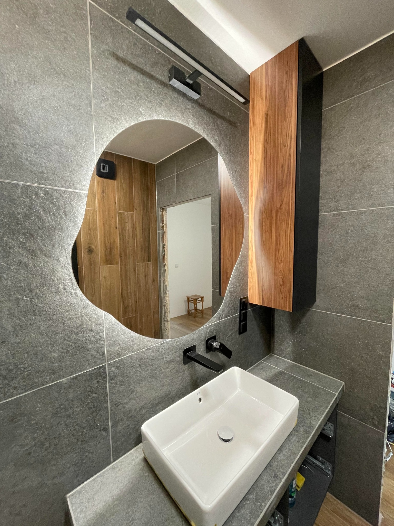 Nowoczesna łazienka z szarymi płytkami, asymetrycznym lustrem z podświetleniem LED, białą prostokątną umywalką i drewnianą szafką wiszącą z czarnymi bokami.