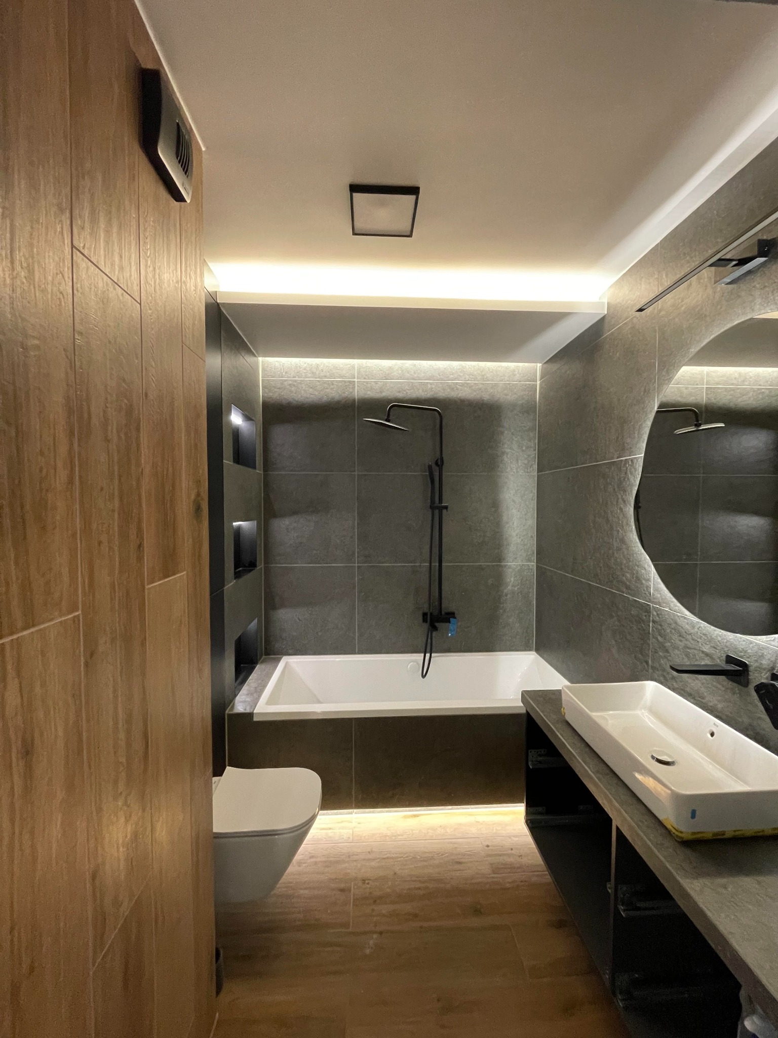Nowoczesna łazienka z szarymi płytkami, podświetleniem LED, asymetrycznym lustrem i drewnopodobną ścianą z ukrytymi wnękami.