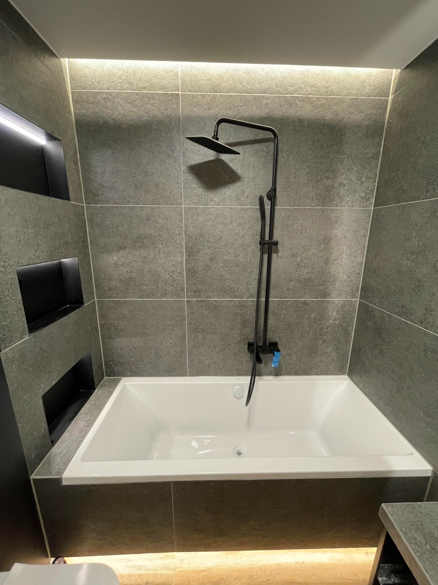 Nowoczesna łazienka z wanną, czarną armaturą i szarymi płytkami z podświetlanymi wnękami.