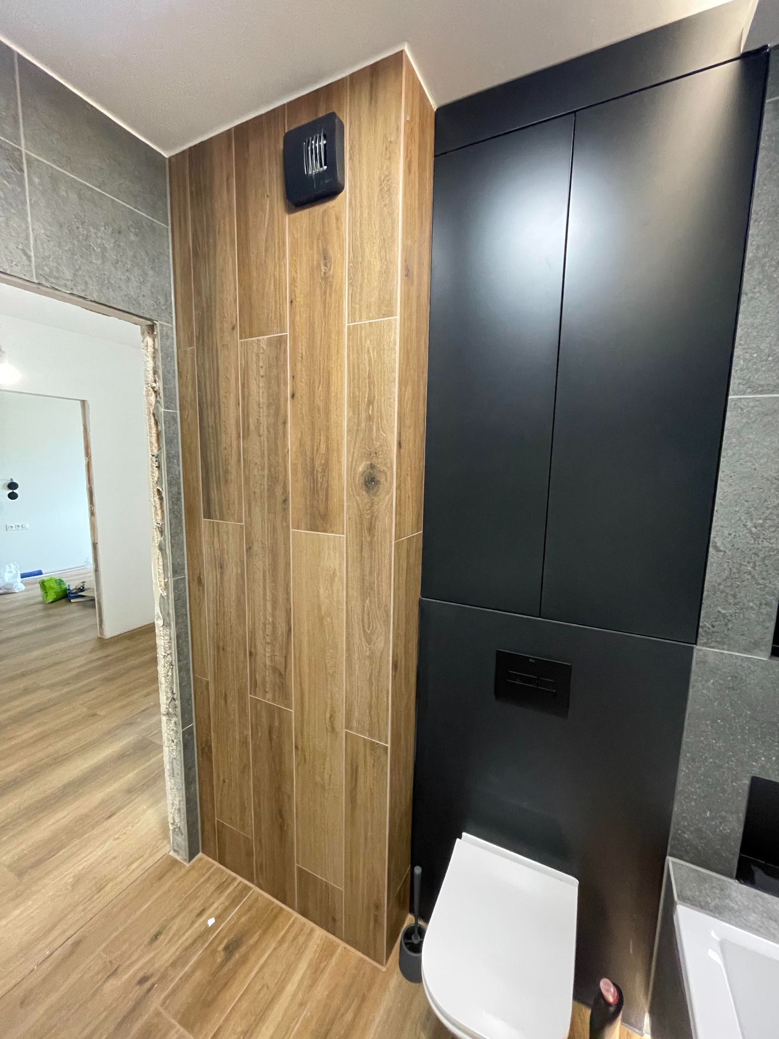 Wykończona łazienka z drewnopodobnymi płytkami na ścianie, czarną zabudową meblową, podwieszaną toaletą i fragmentem umywalki z szarą płytką.