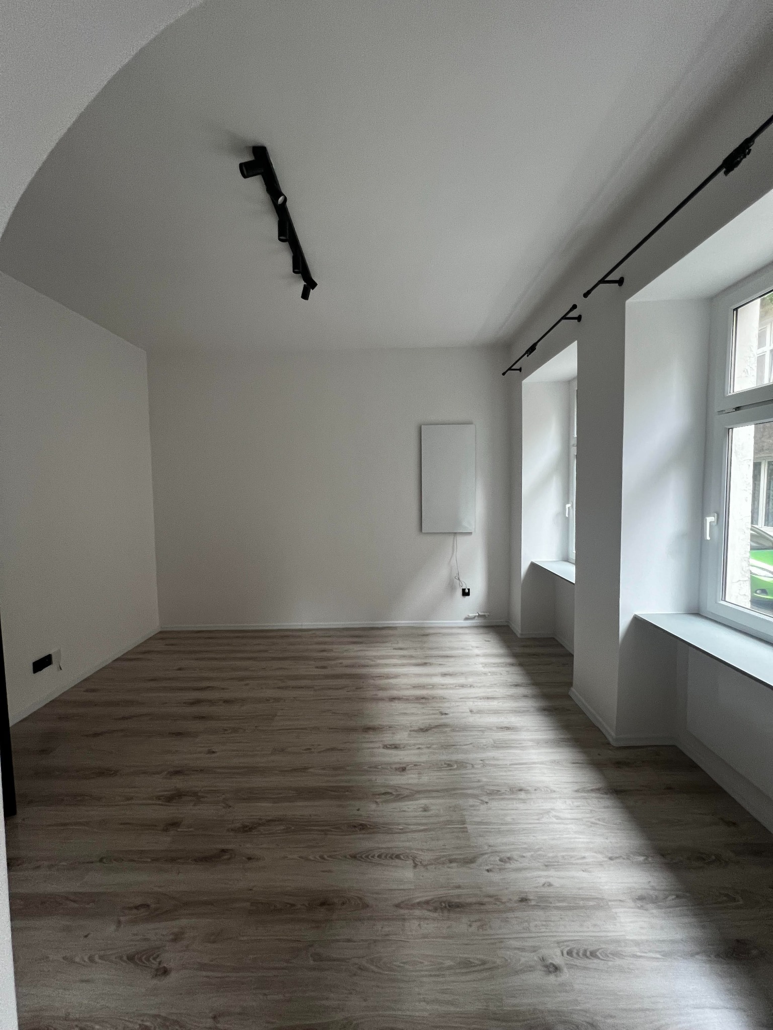 Wnętrze pustego, odnowionego pokoju z panelami podłogowymi imitującymi drewno, białymi ścianami, nowoczesnym oświetleniem szynowym na suficie i grzejnikiem elektrycznym na ścianie.