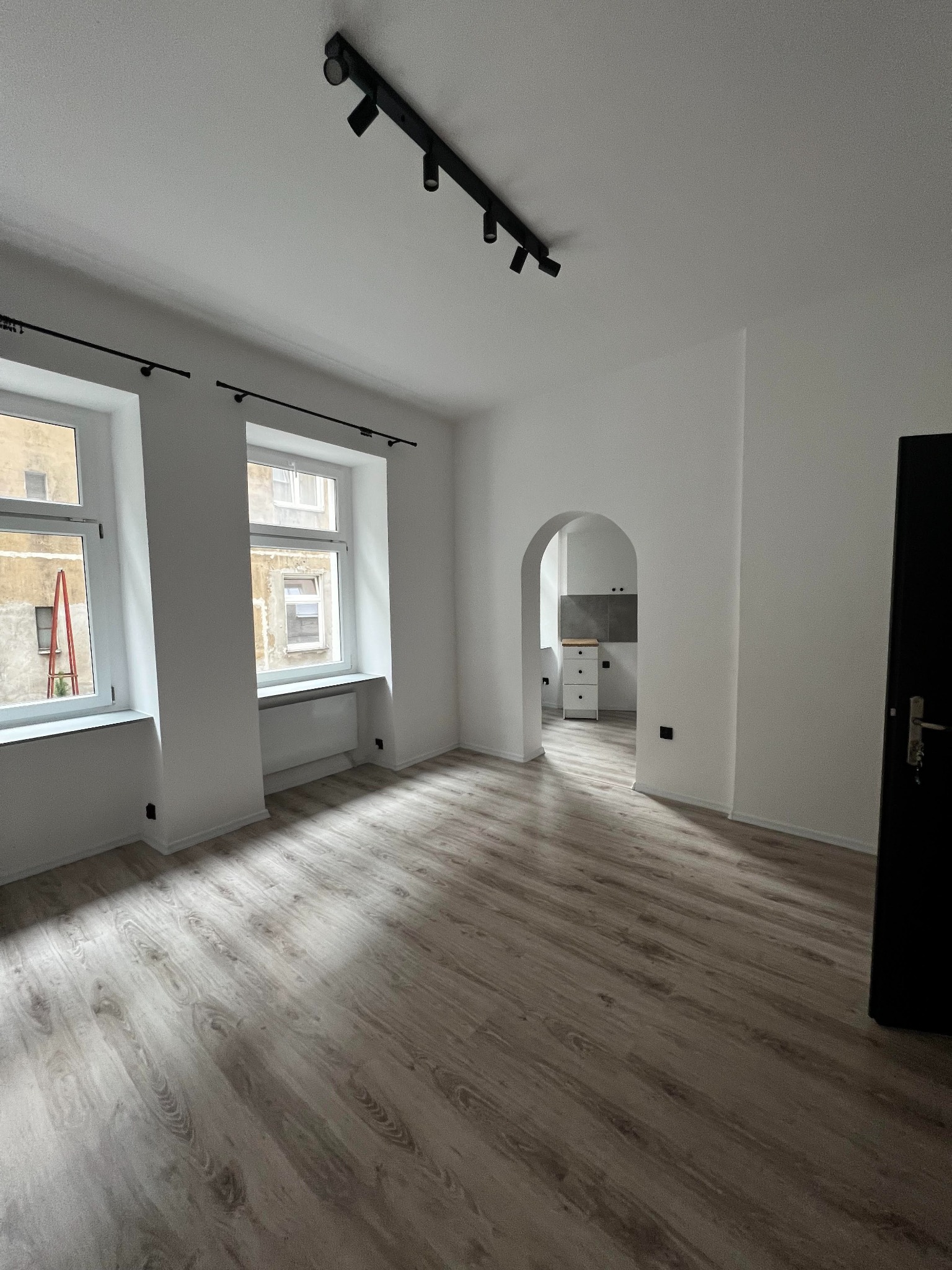 Pusty, świeżo wyremontowany pokój z parkietem, białymi ścianami, dwoma oknami z widokiem na sąsiedni budynek i czarnym oświetleniem szynowym na suficie. W tle widoczne przejście łukowe do kuchni.