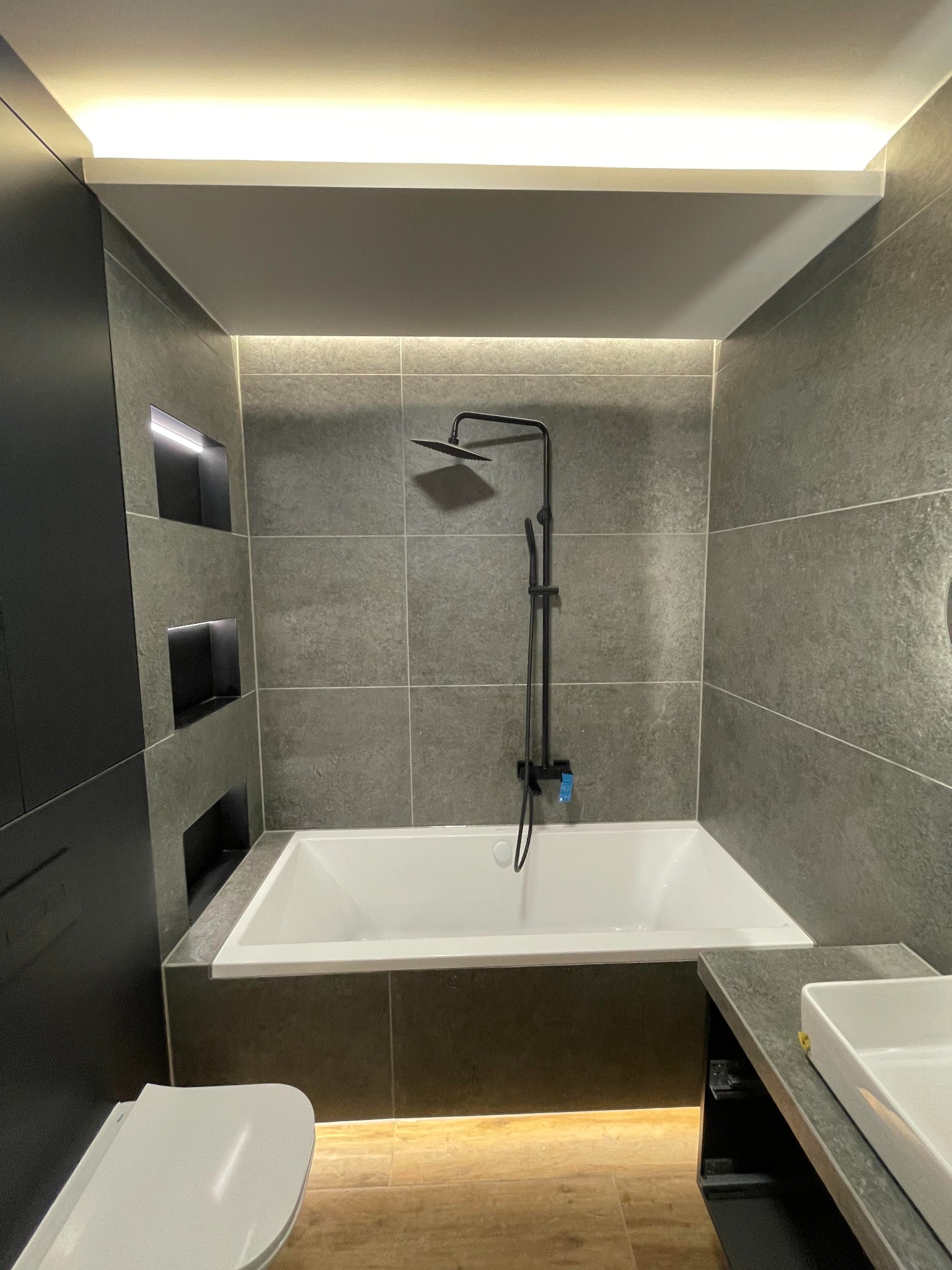 Nowoczesna łazienka w Komornikach z szarymi płytkami, podświetlanymi wnękami i czarną armaturą prysznicową nad białą wanną.