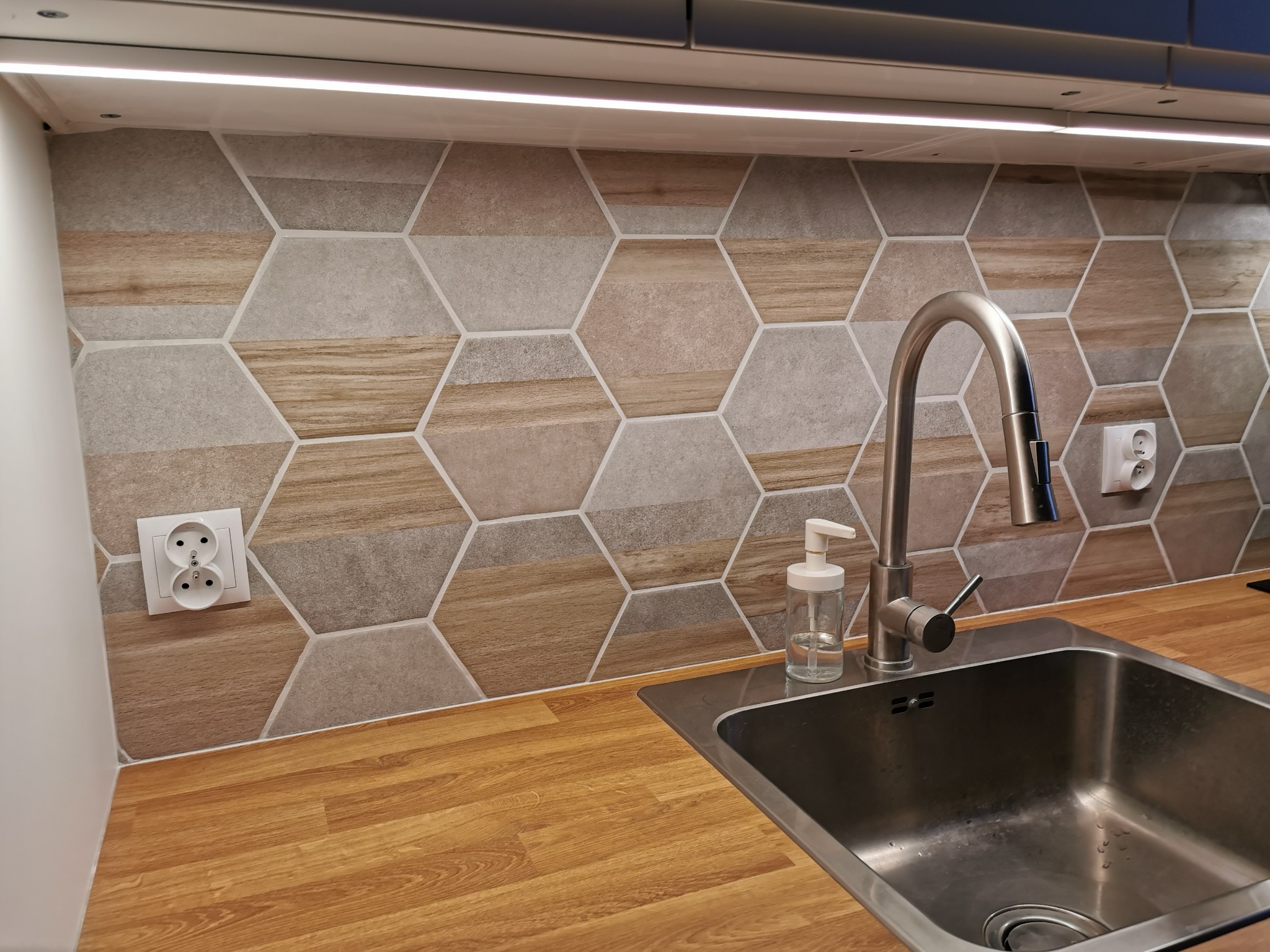 Ściana nad blatem kuchennym wyłożona heksagonalnymi płytkami w odcieniach szarości i drewna, bateria kuchenna z dozownikiem na płyn, drewnopodobny blat oraz gniazdka elektryczne.
