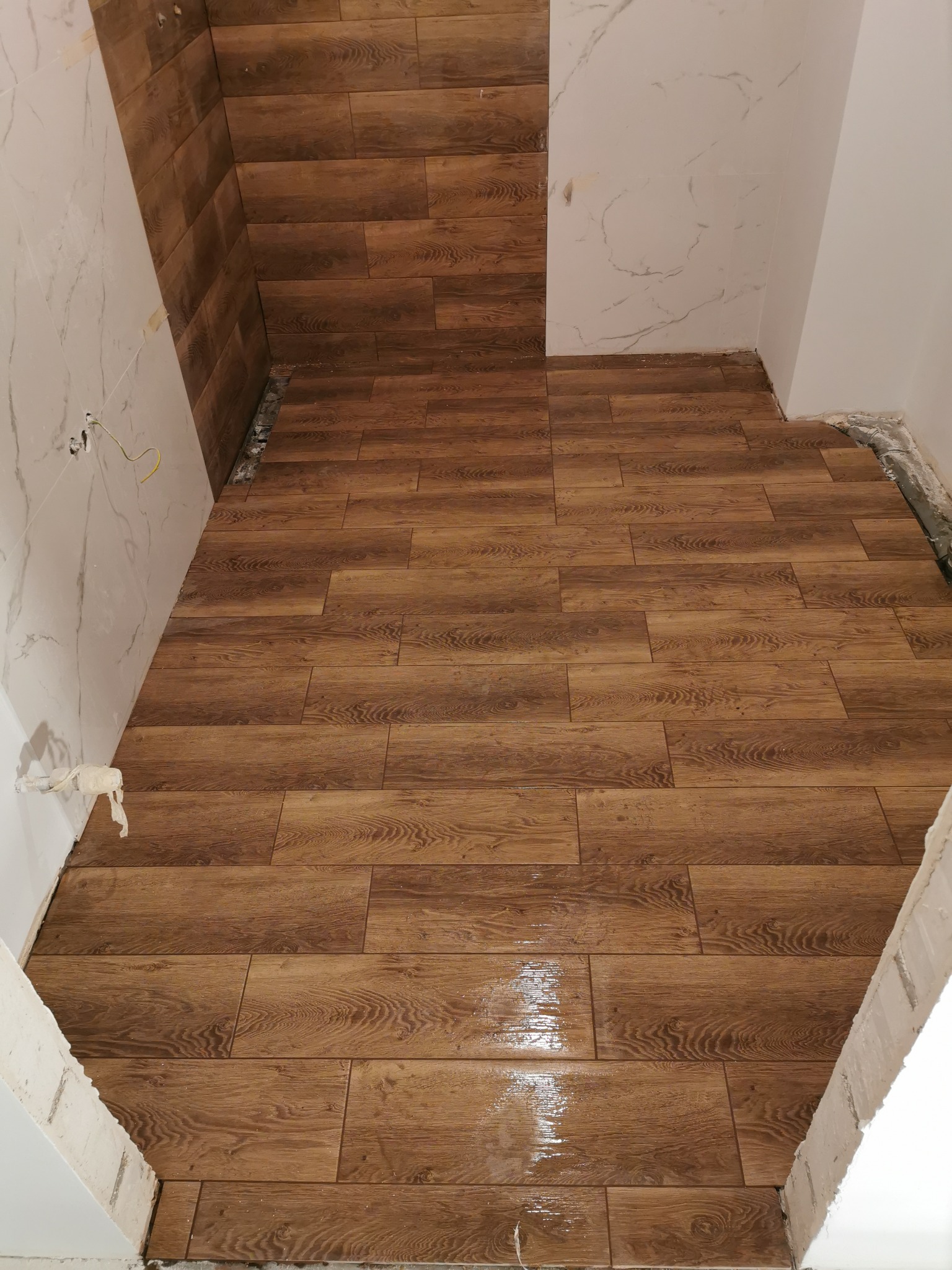 Układanie brązowych płytek imitujących drewno na podłodze i częściowo na ścianie łazienki, widoczne niedokończone fragmenty ścian i wystające przewody.