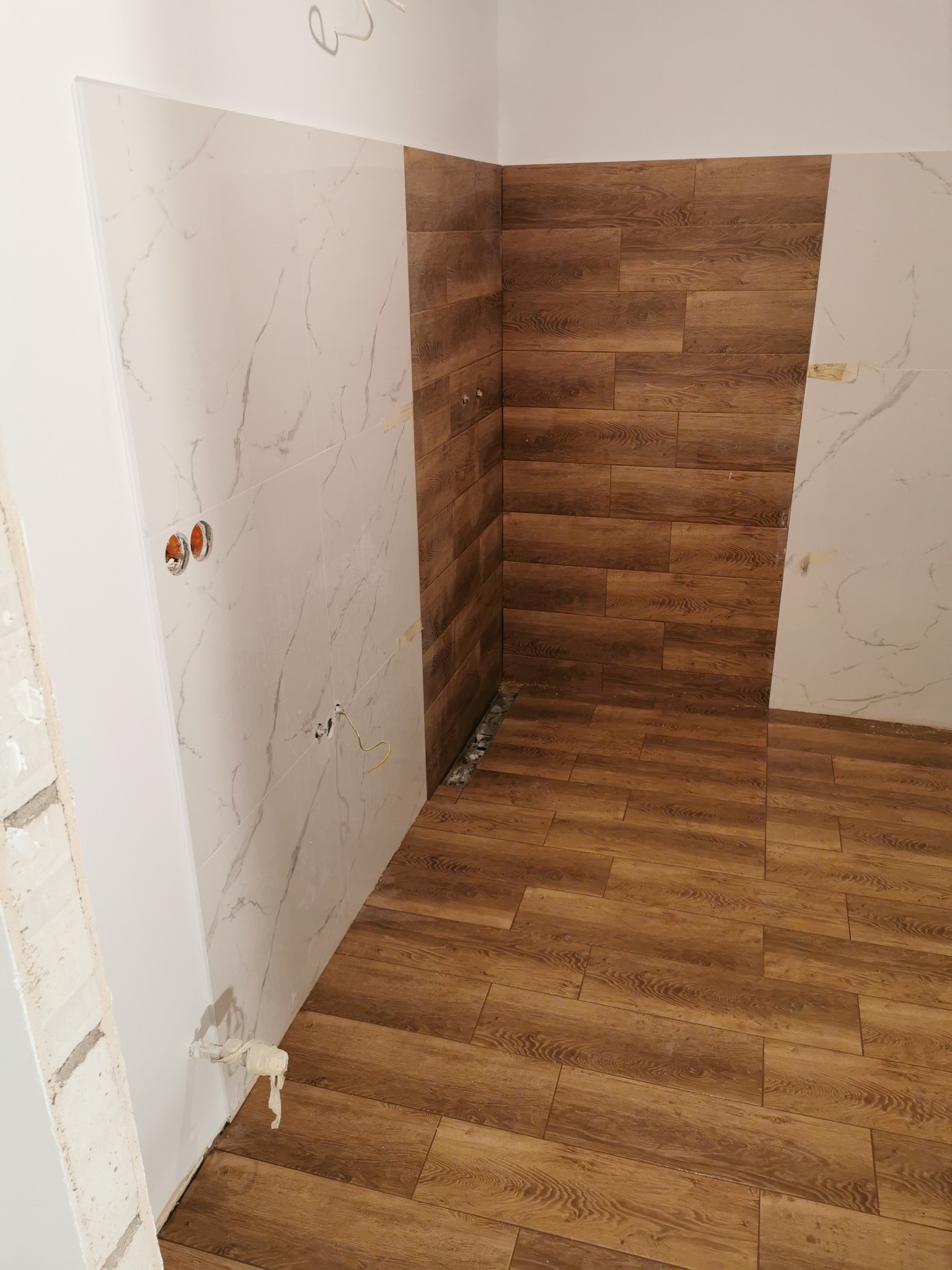 Wykończona łazienka z połączeniem płytek imitujących drewno i marmur, widoczne otwory na gniazdka elektryczne i wyprowadzenia hydrauliczne w ścianie.