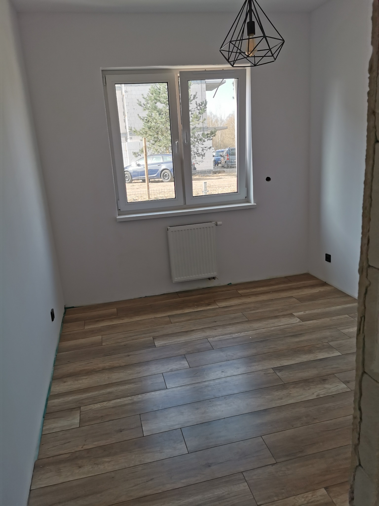Pusty pokój po remoncie z drewnopodobną podłogą, białymi ścianami, grzejnikiem pod oknem i nowoczesną lampą wiszącą.