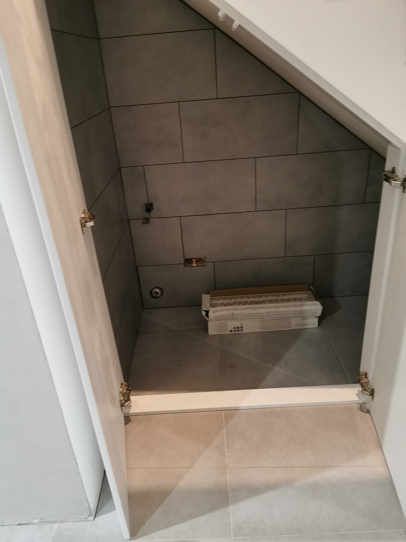 Wnęka pod schodami wyłożona szarymi płytkami ceramicznymi, widoczne przyłącza wodne i kanalizacyjne, karton z płytkami na podłodze, otwarte białe drzwi.