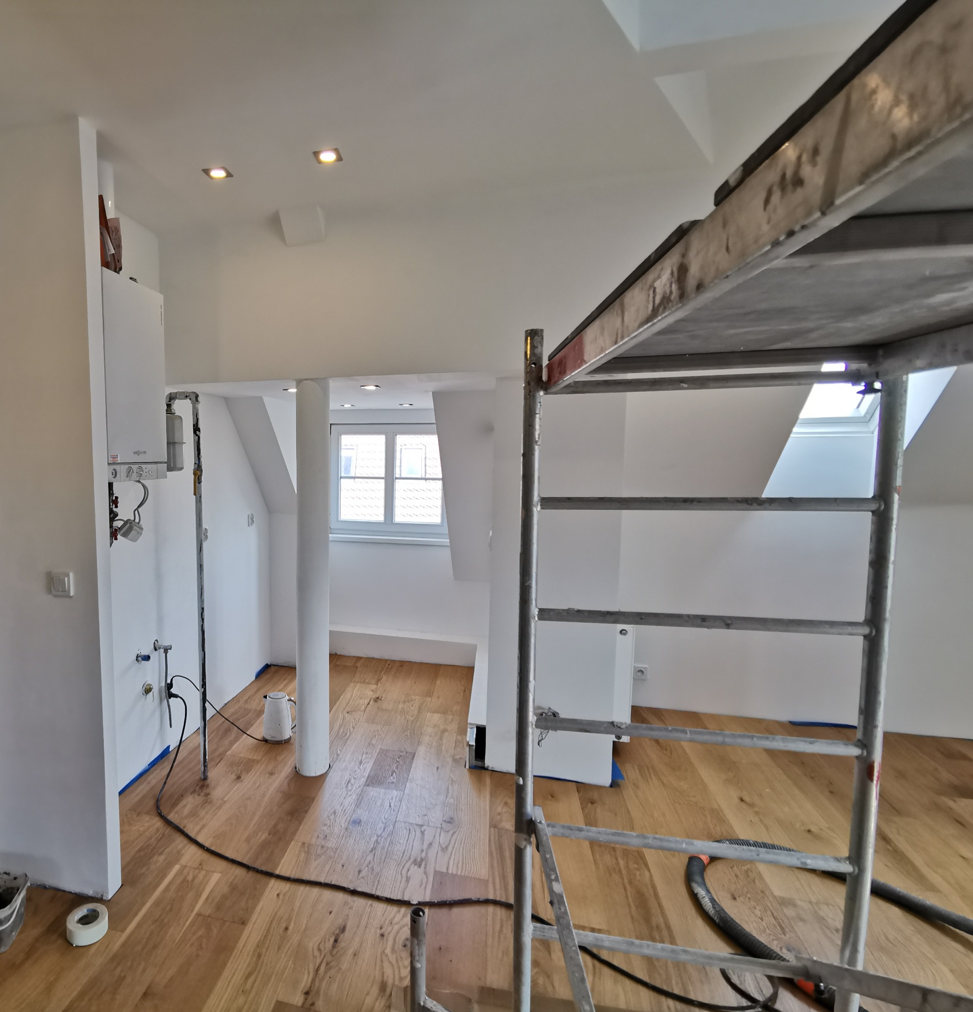 Wnętrze poddasza w trakcie remontu, widoczny piec gazowy, drewniana podłoga zabezpieczona taśmą malarską, kolumna, okno dachowe i metalowy ruszt.