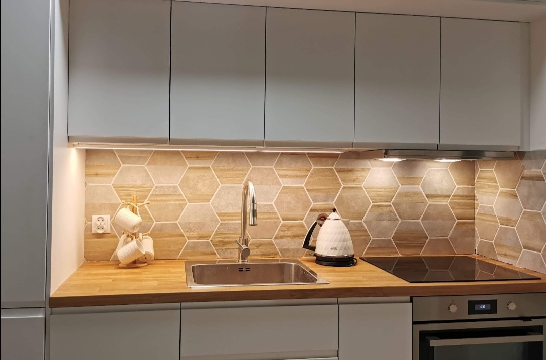 Nowoczesna kuchnia z heksagonalnymi płytkami imitującymi drewno, oświetleniem LED pod szafkami i drewnianym blatem, na którym stoją czajnik elektryczny i suszarka na kubki.