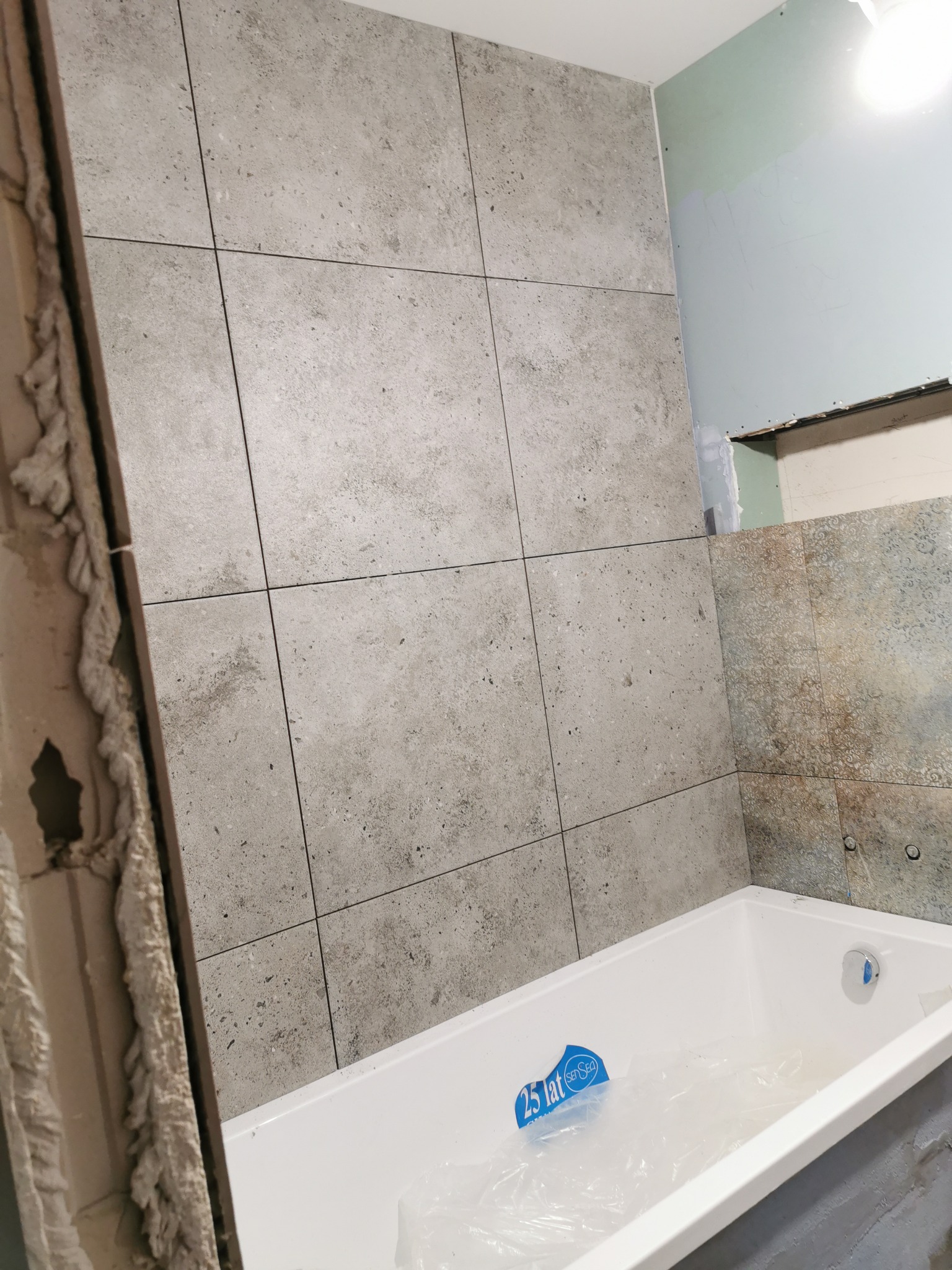 Wnętrze łazienki w trakcie remontu: biała wanna zabezpieczona folią, ściana wyłożona szarymi, kwadratowymi płytkami imitującymi kamień, surowa ściana z zielonymi fragmentami i odsłoniętą konstrukcją.