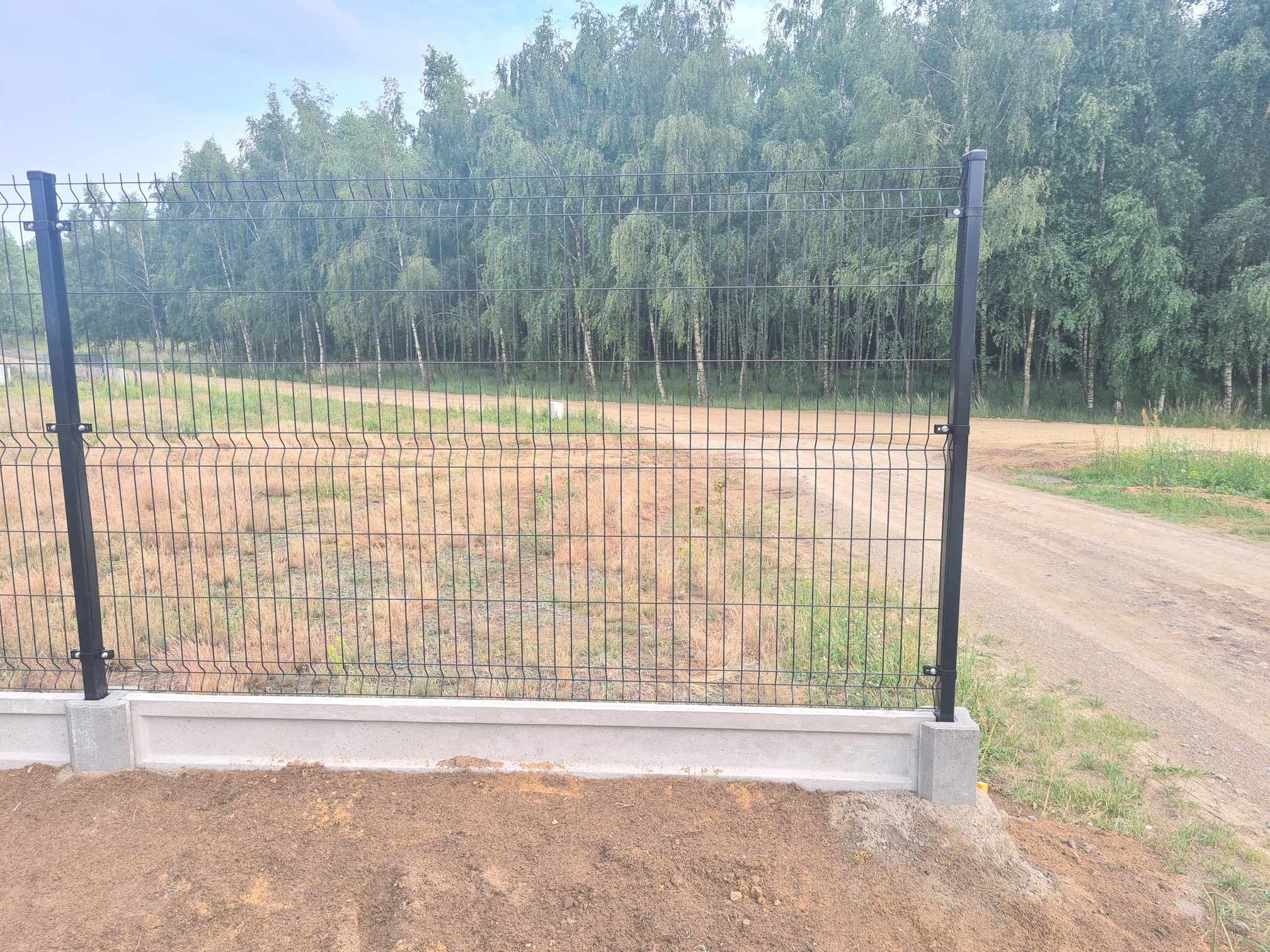 Czarna siatka ogrodzeniowa panelowa na betonowej podmurówce, widok z bliska na tle drzew i drogi gruntowej.