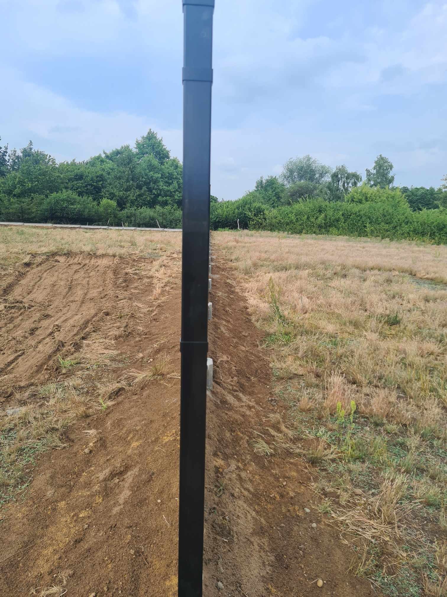 Fragment ogrodzenia panelowego w trakcie montażu, widoczne słupki i części paneli w surowym krajobrazie z przygotowanym terenem pod budowę.