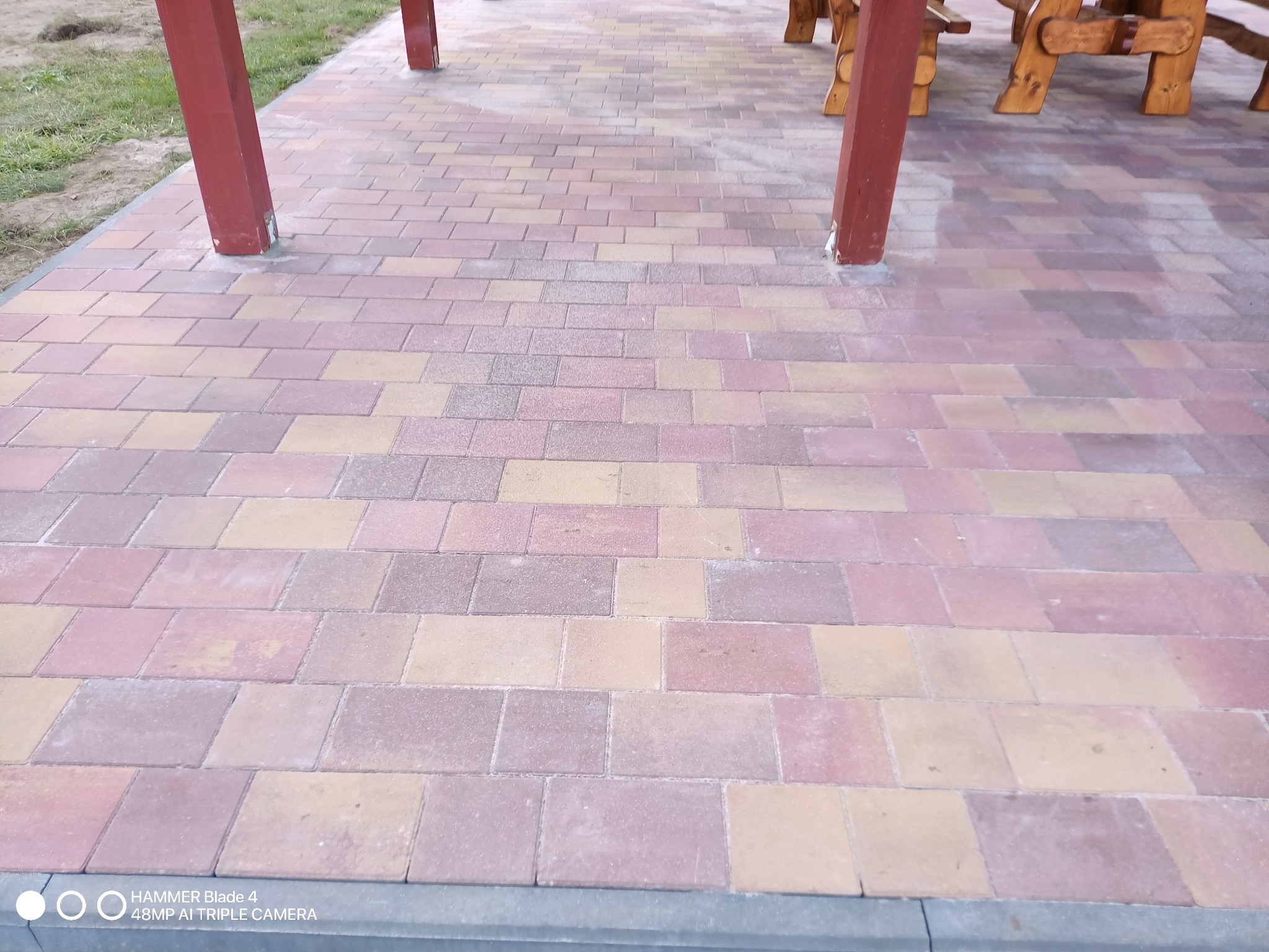 Kolorowa kostka brukowa ułożona pod zadaszeniem, widoczne podpory konstrukcji i fragment drewnianego stołu.