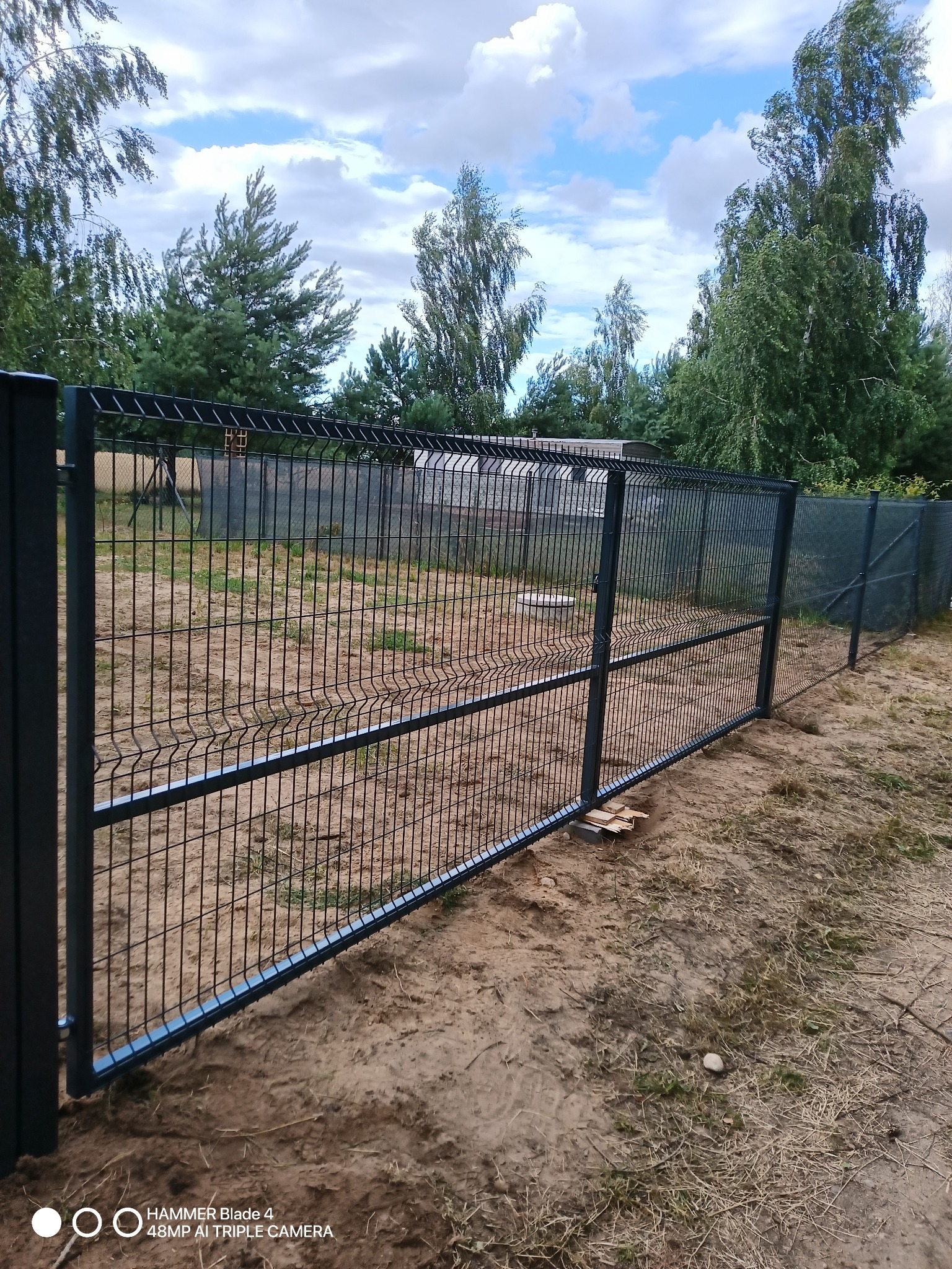 Ciemnoszare ogrodzenie panelowe z metalowej siatki na piaszczystym terenie, widok na drzewa i błękitne niebo z chmurami.