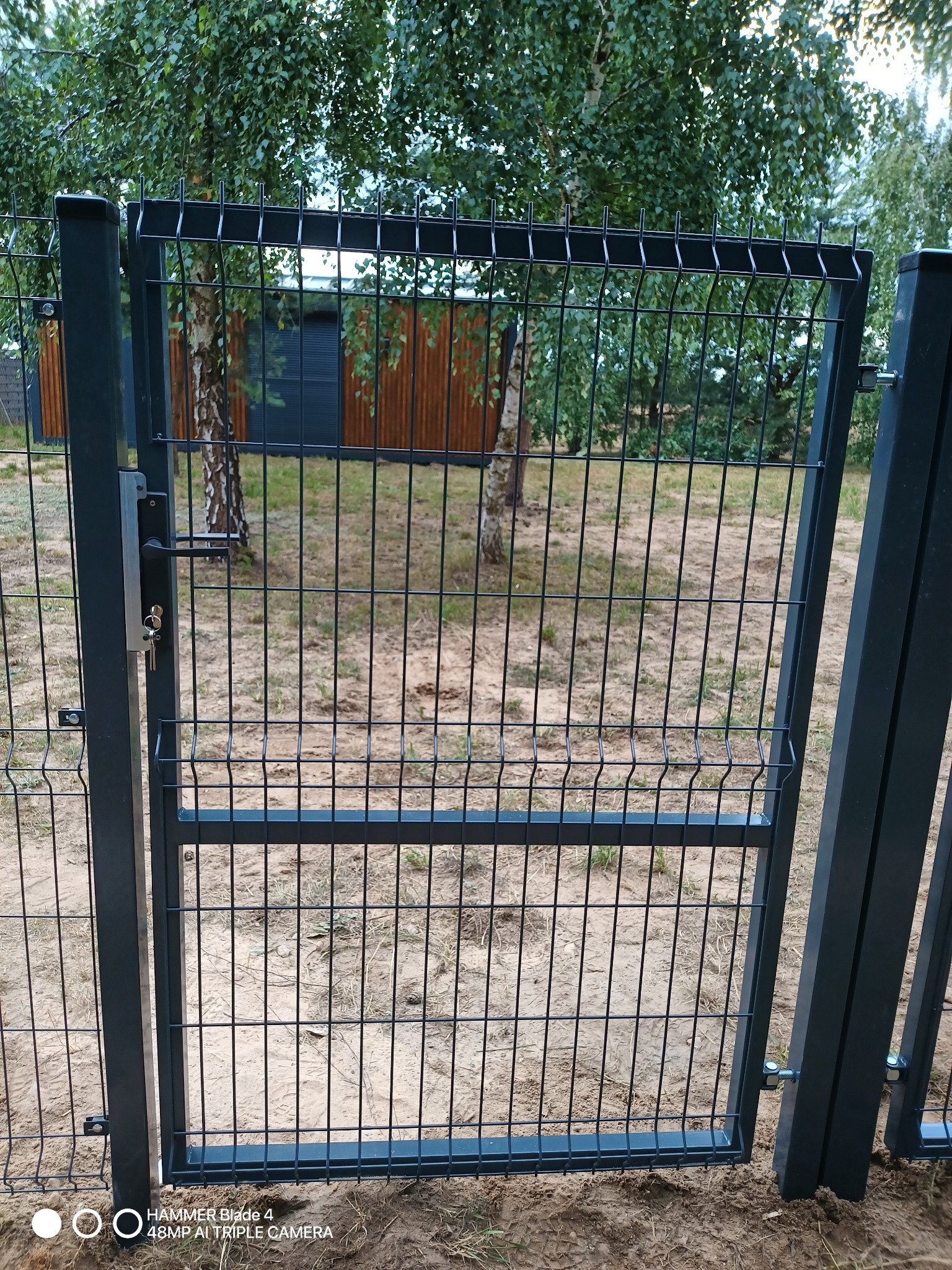 Nowa, pomalowana na grafitowo, metalowa furtka panelowa z kluczem w zamku, zamontowana w ogrodzeniu na piaszczystym terenie, z widocznym budynkiem gospodarczym w tle.