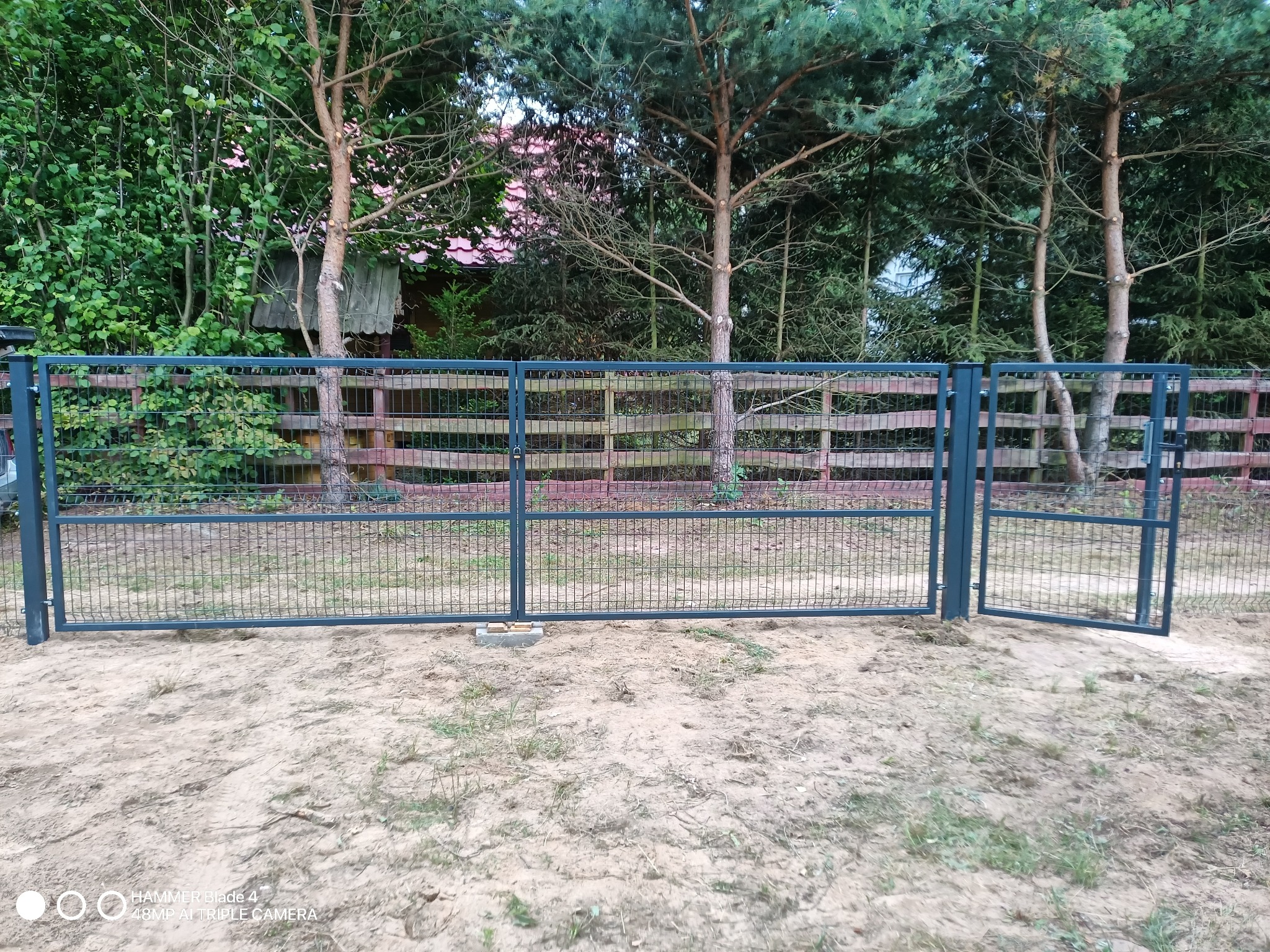 Zamontowana brama wjazdowa z siatki w metalowej ramie w Lubieniu Kujawskim, z widoczną otwartą furtką i surowym, piaszczystym podłożem.