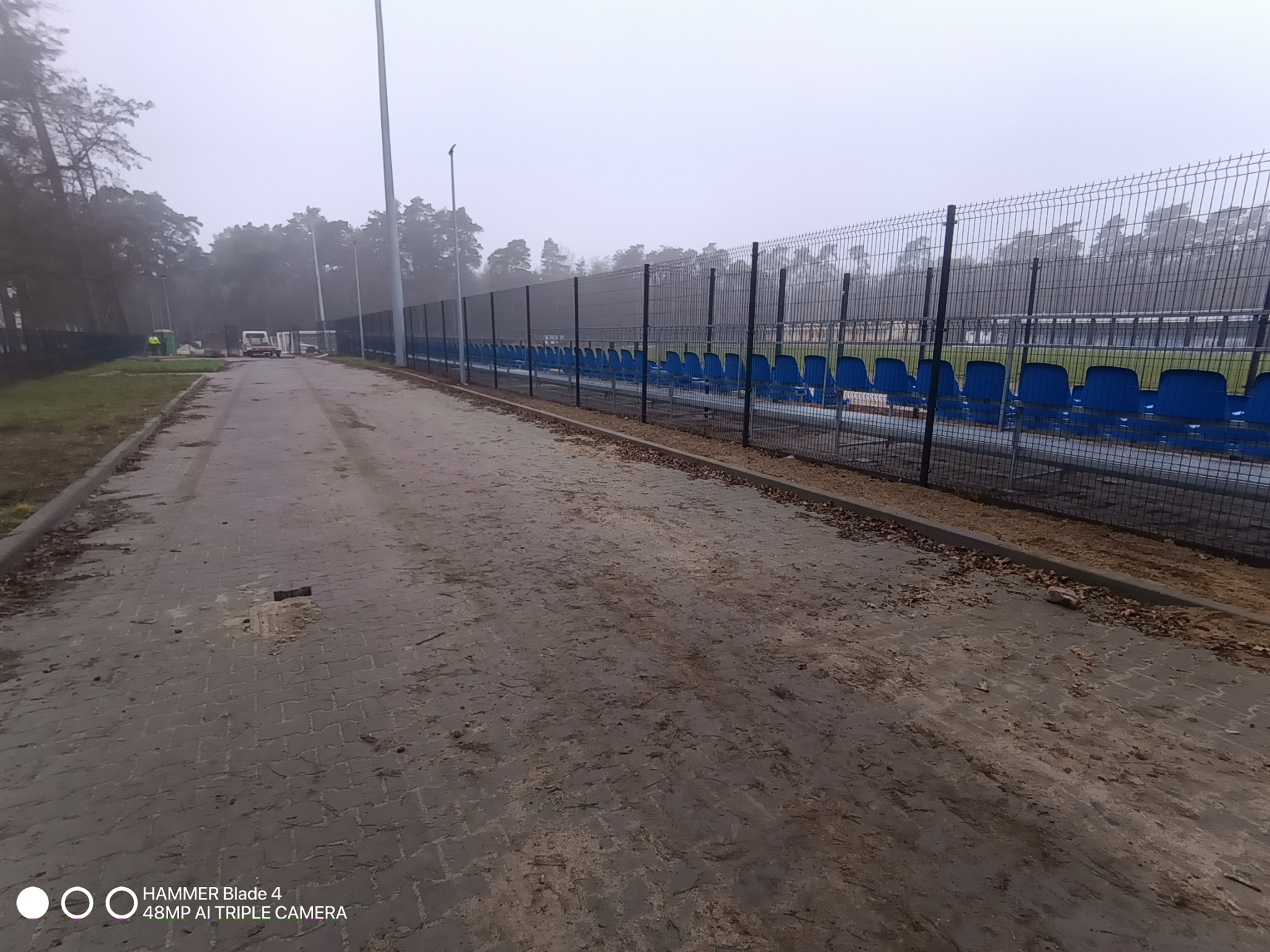 Długie, metalowe ogrodzenie z siatki na stadionie, biegnące wzdłuż rzędu niebieskich krzesełek. Mglista pogoda, widoczna droga z kostki brukowej.