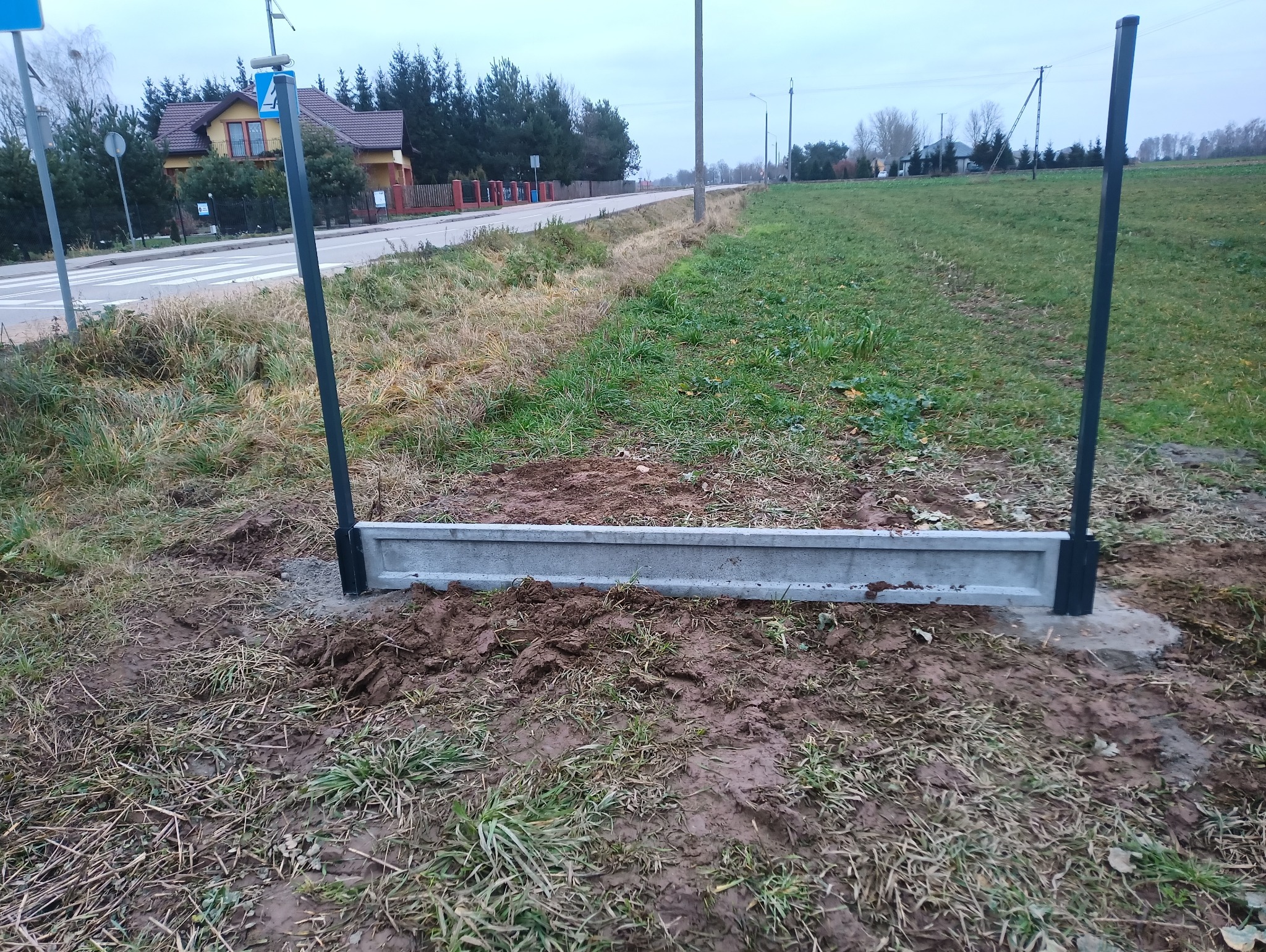 Fragment montażu ogrodzenia metalowego z podmurówką betonową na tle wiejskiego krajobrazu i drogi. Widoczne słupki i element betonowy w ziemi.