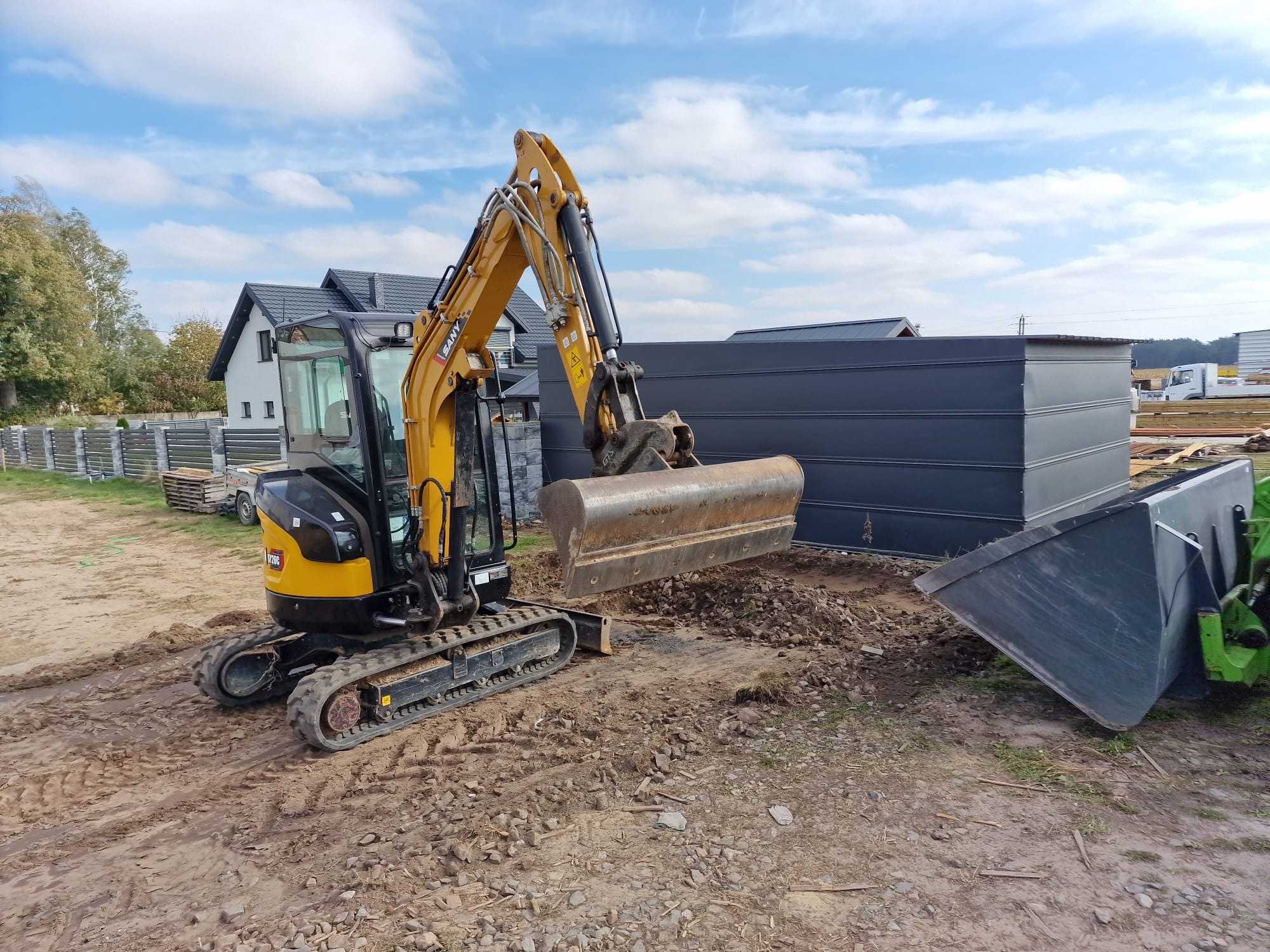 Żółto-czarna mini koparka SANY na placu budowy, z widocznymi śladami opon w ziemi, w tle domy i niebieskie niebo z chmurami.